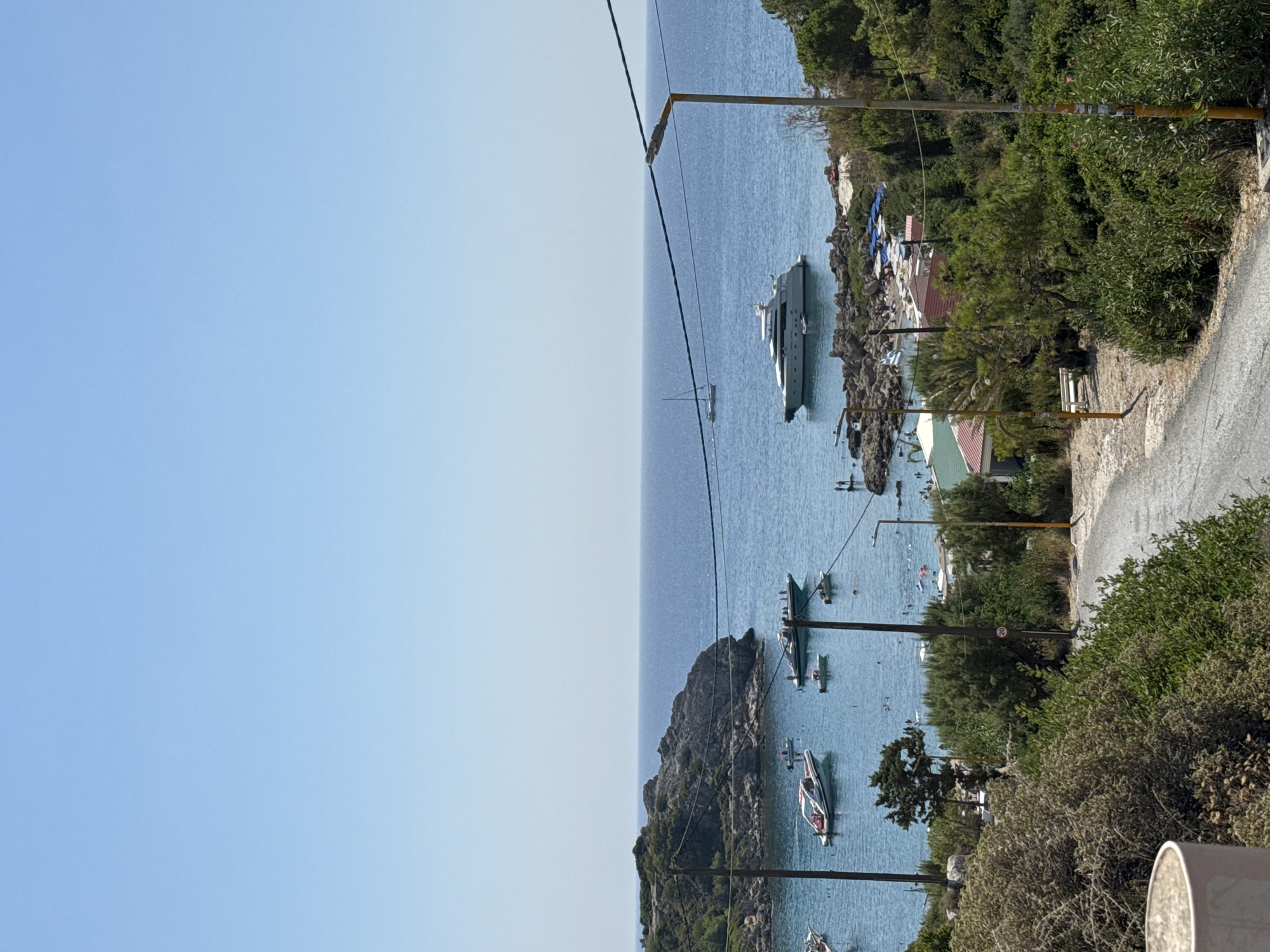 The image depicts a scenic coastal view in Rhodes, Greece, likely from a vantage point overlooking a bay or harbor. The sky is a clear, bright blue, transitioning to a slightly lighter hue near the horizon. The sea is a deep blue, calm with gentle waves, dotted with various boats. In the foreground, a paved road curves to the right, bordered by lush green foliage and trees. There are also two utility poles with wires running across the frame.
Several boats are visible in the water. Closest to the shore in the middle ground, there are several smaller boats, some appearing to be speedboats or dinghies, anchored or moving slowly. Further out, a large, white luxury yacht is prominently featured, with a smaller dinghy tethered to its stern. A lone sailboat is visible in the distance, its mast a thin line against the horizon.
On the rocky shoreline and a small beach area, there are signs of human activity. People are seen scattered along the beach and near the water, some appearing to be swimming or relaxing under umbrellas. Small buildings with red roofs are nestled amongst the trees on the shore. A Greek flag is flying on a rocky outcrop on the right side of the image. The overall impression is a vibrant, sunny day in a popular coastal tourist destination.