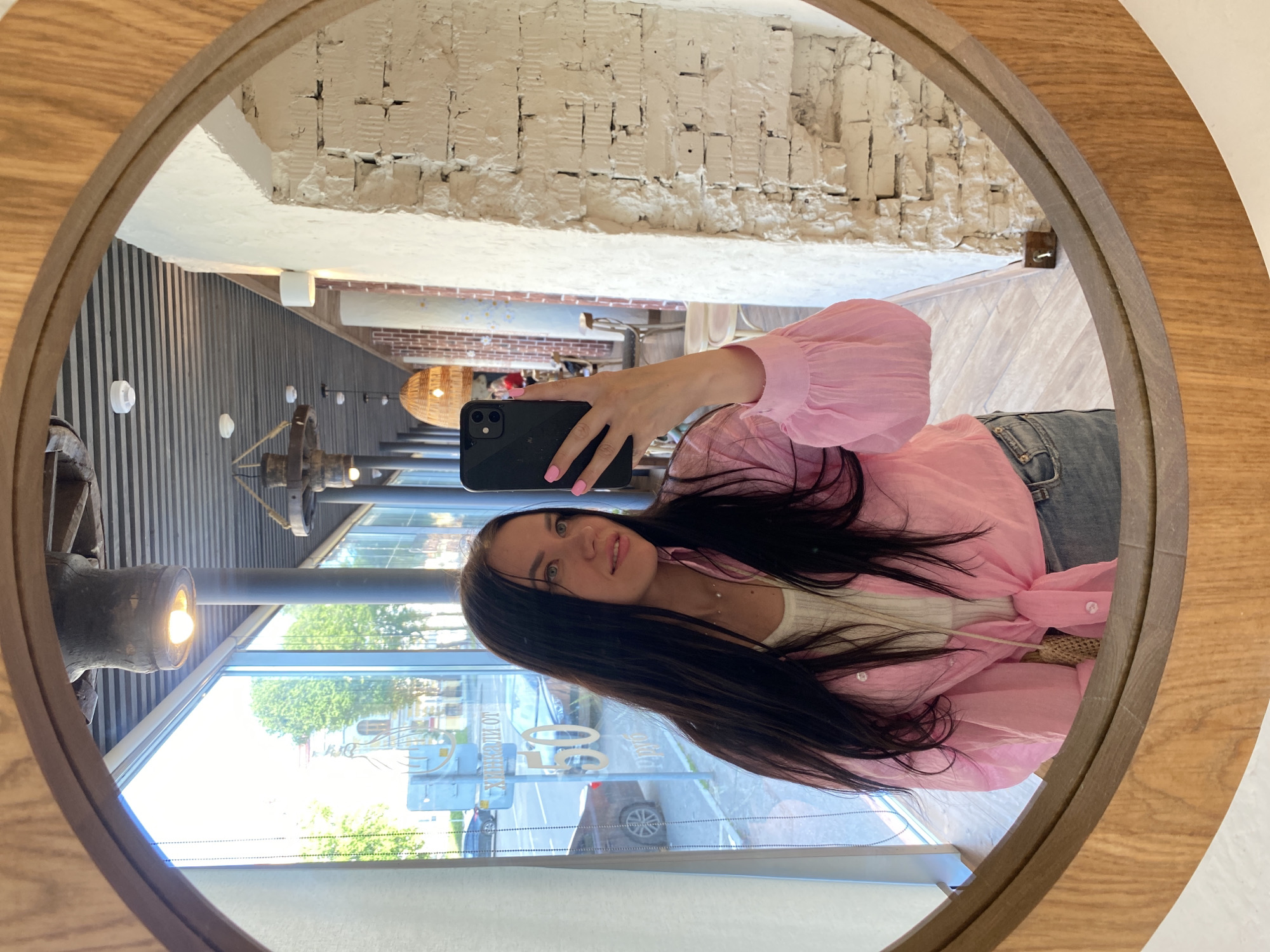 In a circular mirror with a wooden frame, a young woman with long dark hair takes a selfie. She is wearing a pink oversized shirt tied at the waist over a white top and blue jeans. The mirror reflects a sunlit interior space with large windows, exposed brick walls, and wooden floors. Outside the windows, a street scene in Perm, Russia, is visible, with cars and buildings. The interior appears to be a cafe or restaurant, with pendant lights and industrial-style decor. The woman is holding a black smartphone to take the photo.