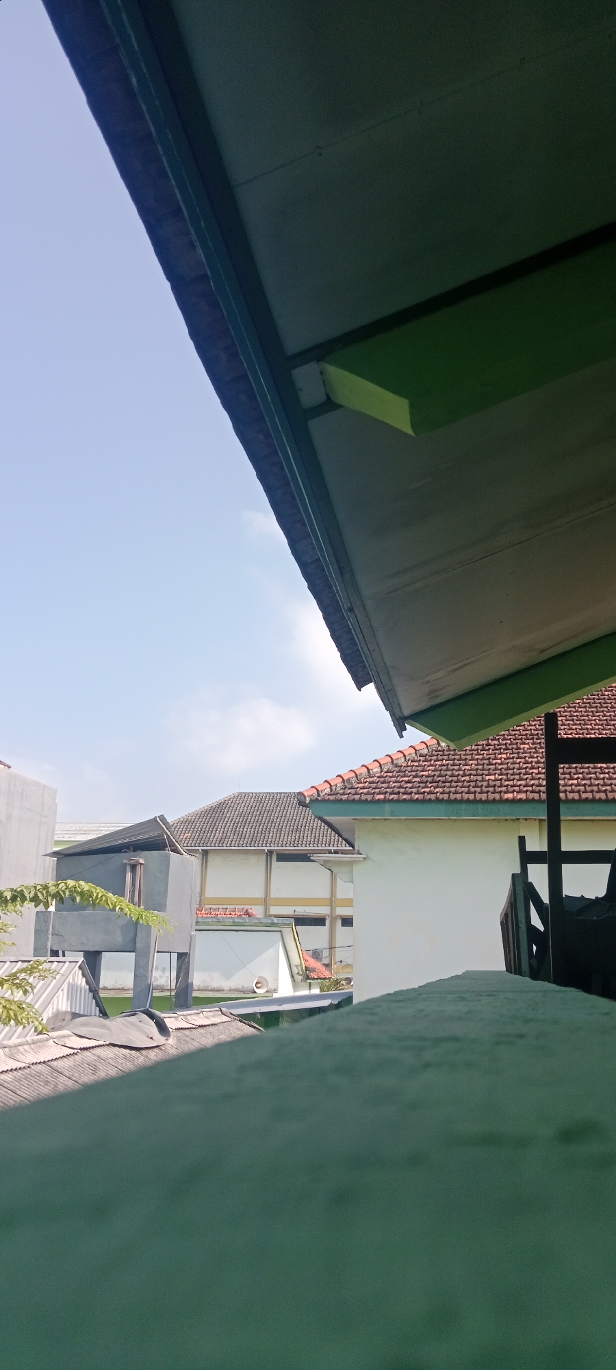 The image depicts an outdoor scene with several buildings under a bright, clear sky with scattered clouds. In the foreground, a green surface, possibly a wall or railing, is covered with a light dusting of what appears to be sand or fine debris. Above this, the underside of a roof structure with green trim and white paneling is visible, angled upwards towards the sky.
Further back, several buildings are visible. To the left, a light gray, modern building with visible structural elements. To its right, a building with a gray tiled roof and a white exterior wall with yellow accents. A smaller structure with a flat, dark roof and concrete supports is situated in front of this building. To the far right, a white building with a red tiled roof is partially visible, with a black wooden structure in the foreground.
The scene suggests an urban or suburban environment. Based on visual cues and common architectural styles in Indonesia, this scene could be located in the city of Tuban, Indonesia, potentially in a residential or institutional area. No people or specific actions are depicted in the image. The environment is characterized by daylight and clear weather.