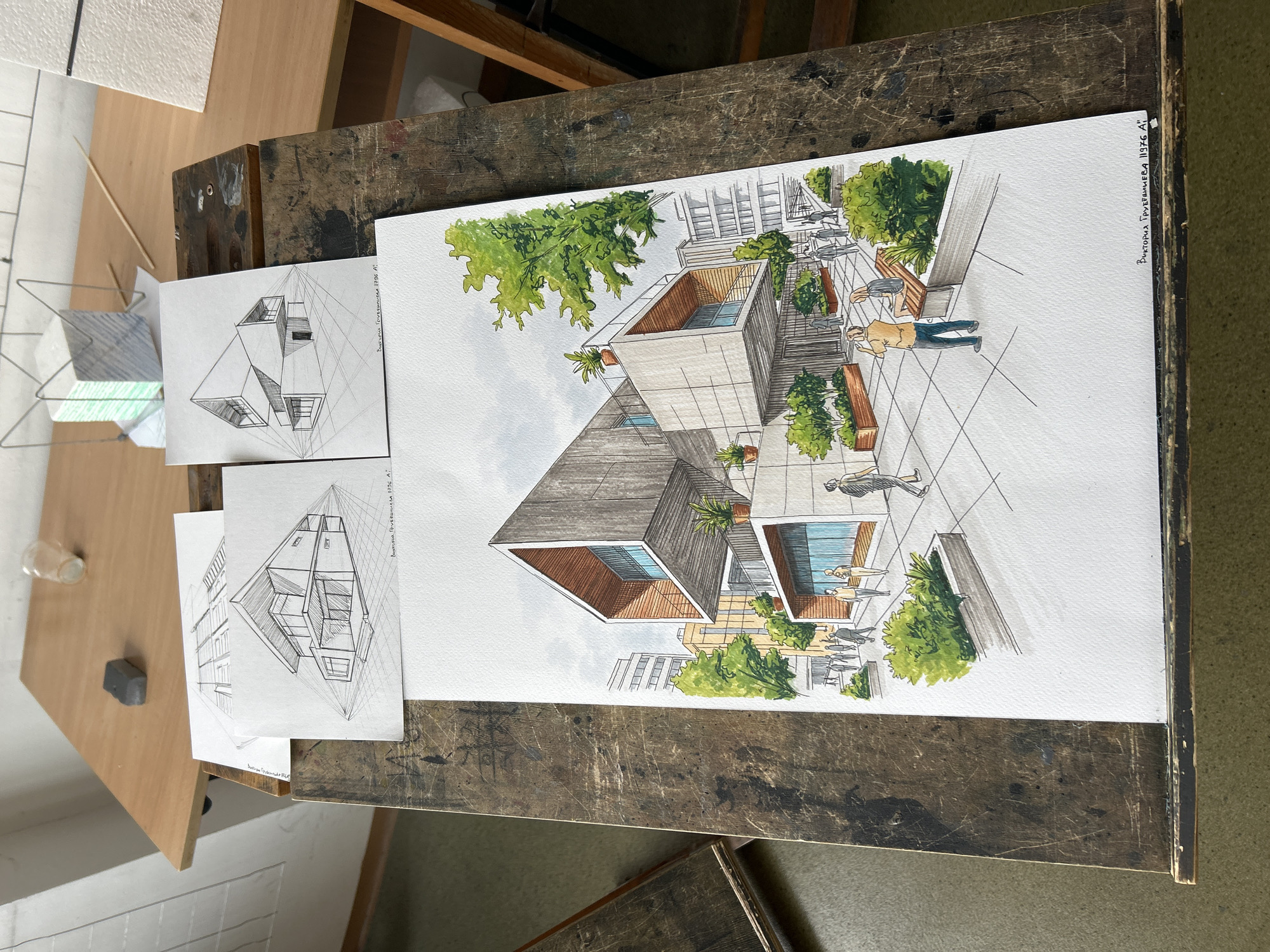 The image shows a desk with architectural drawings. The main drawing is a colored perspective rendering of a modern building with a distinctive angled roof. The building is surrounded by trees and appears to be situated in an urban environment, evidenced by other buildings in the background. People are depicted walking and sitting in the foreground, suggesting a public plaza or outdoor area.
Two smaller, black and white perspective drawings of the same building, possibly sketches or initial designs, lie to the left of the colored rendering. These drawings include vanishing point lines, indicating a focus on perspective.
The drawings are placed on a wooden desk with a worn, textured surface, suggesting a workspace. Based on the writing "Виктория Трухмилева 11936 А1" on the right side of the colored rendering, it could be presumed that this is a drawing done by Victoria Trukhmileva. The location of the scene in the city of Sofia, Bulgaria cannot be determined from the image.