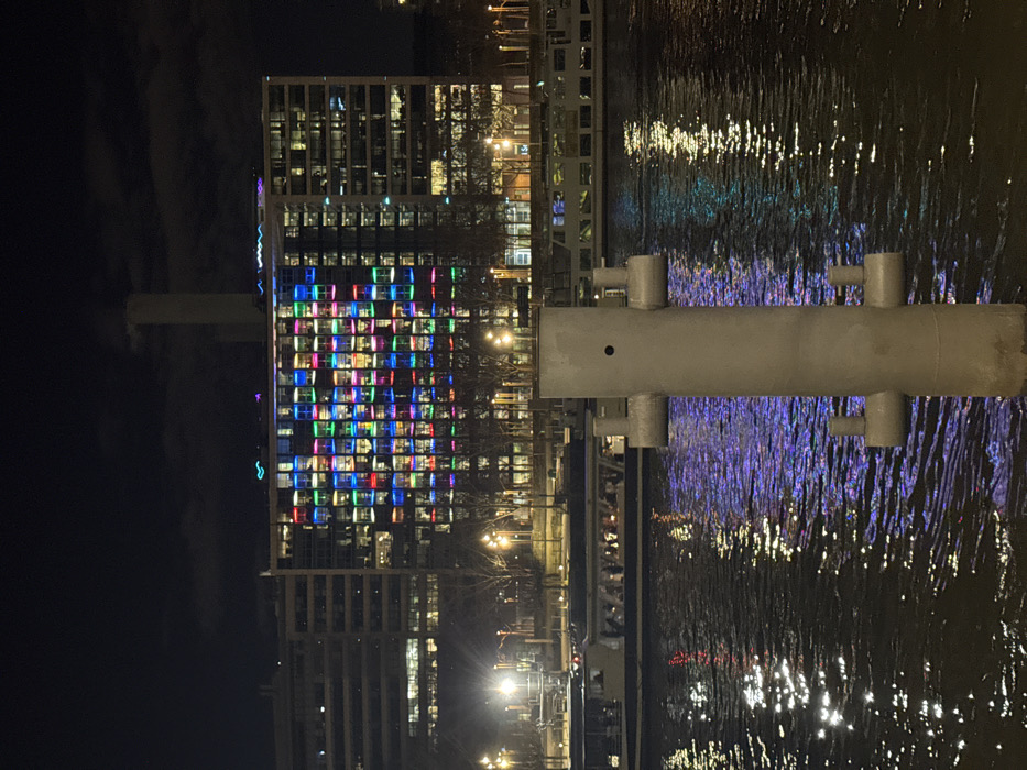 Night scene of a modern building with colorful lights in a grid pattern reflecting in the water. A section of a bridge or pier, appearing cylindrical with attached pipes, is visible in the foreground. The water shows blurred reflections of the building and nearby lights, creating a shimmering effect. The sky is dark, and the overall ambiance is urban and vibrant.