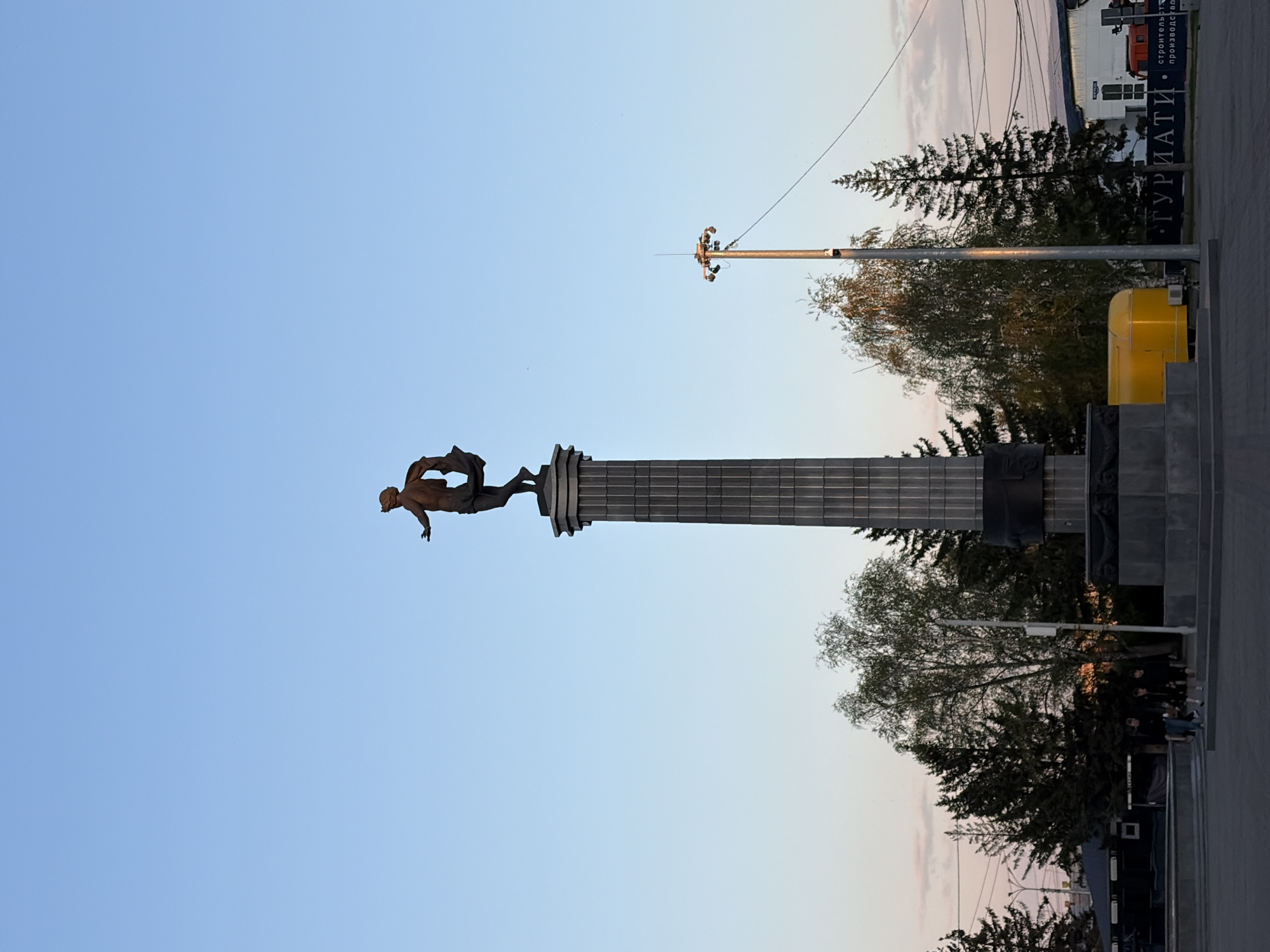 A statue of a muscular man atop a column stands against a clear blue sky. The column is gray and fluted. A tall light pole with several lights and cameras stands adjacent to the statue. Trees surround the base of the monument, and a yellow utility box is visible. A building with signage in Cyrillic can be seen in the background. The statue is located in Krasnoyarsk, Russia.