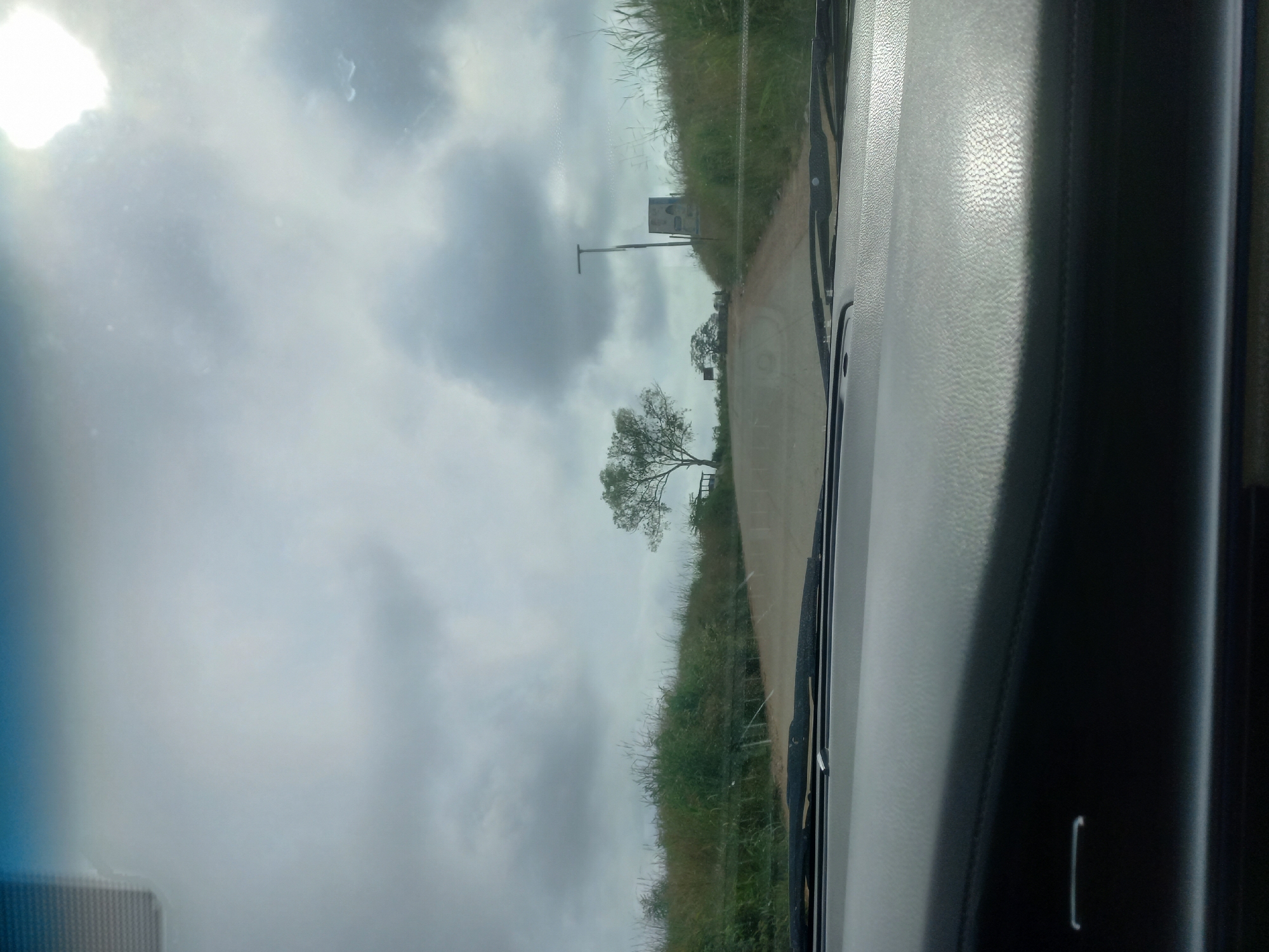 The image captures a view from inside a vehicle, looking out through the windshield. The scene depicts a dirt road ascending a gentle incline, flanked by tall, dry grasses and sparse trees. The sky is heavily overcast with dramatic clouds, partially obscuring a bright sun. On the right side of the road, atop the hill, stands a unique, cylindrical structure with a light fixture extending from its top, and to its left, a solitary tree with sparse foliage. A smaller, similar tree is visible further down the road on the left. The foreground is dominated by the interior of the car, showing the top of the dashboard and the windshield wipers. The overall atmosphere is one of a rural or semi-rural setting under a cloudy, possibly humid, sky. No people are visible in the image.