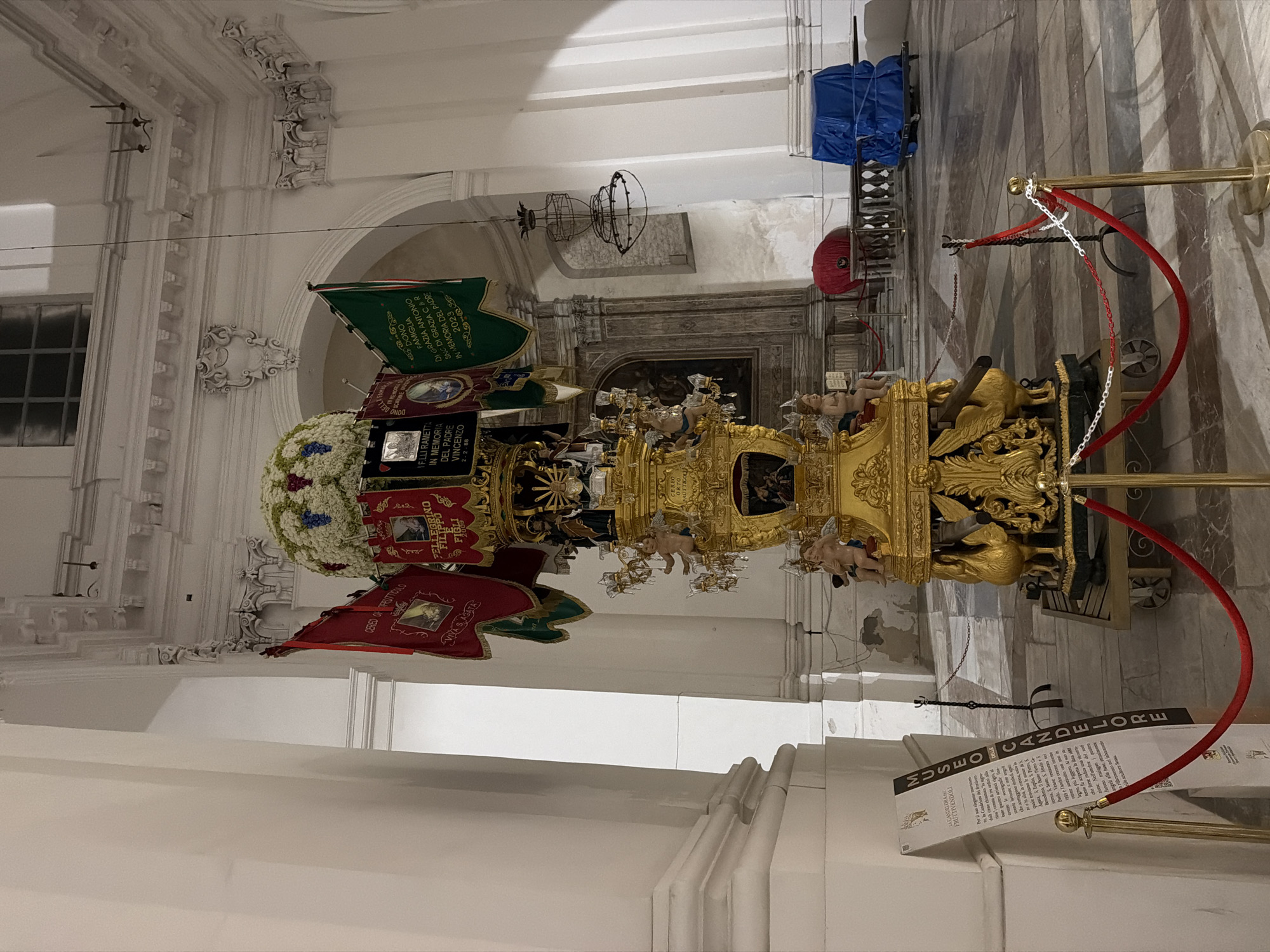 This indoor shot captures a richly ornamented, golden religious structure, potentially a "Candelora", which is on display at the Museo Candelore in Catania, Italy. The Candelora is adorned with banners, flags, and a floral arrangement, and sits atop a wheeled platform. The structure is roped off with red velvet ropes. The architectural setting is ornate with decorative moldings and white walls, while a dark painting or mosaic is visible further in the background. A window is seen on the left. A museum display card stands in the foreground with printed information.