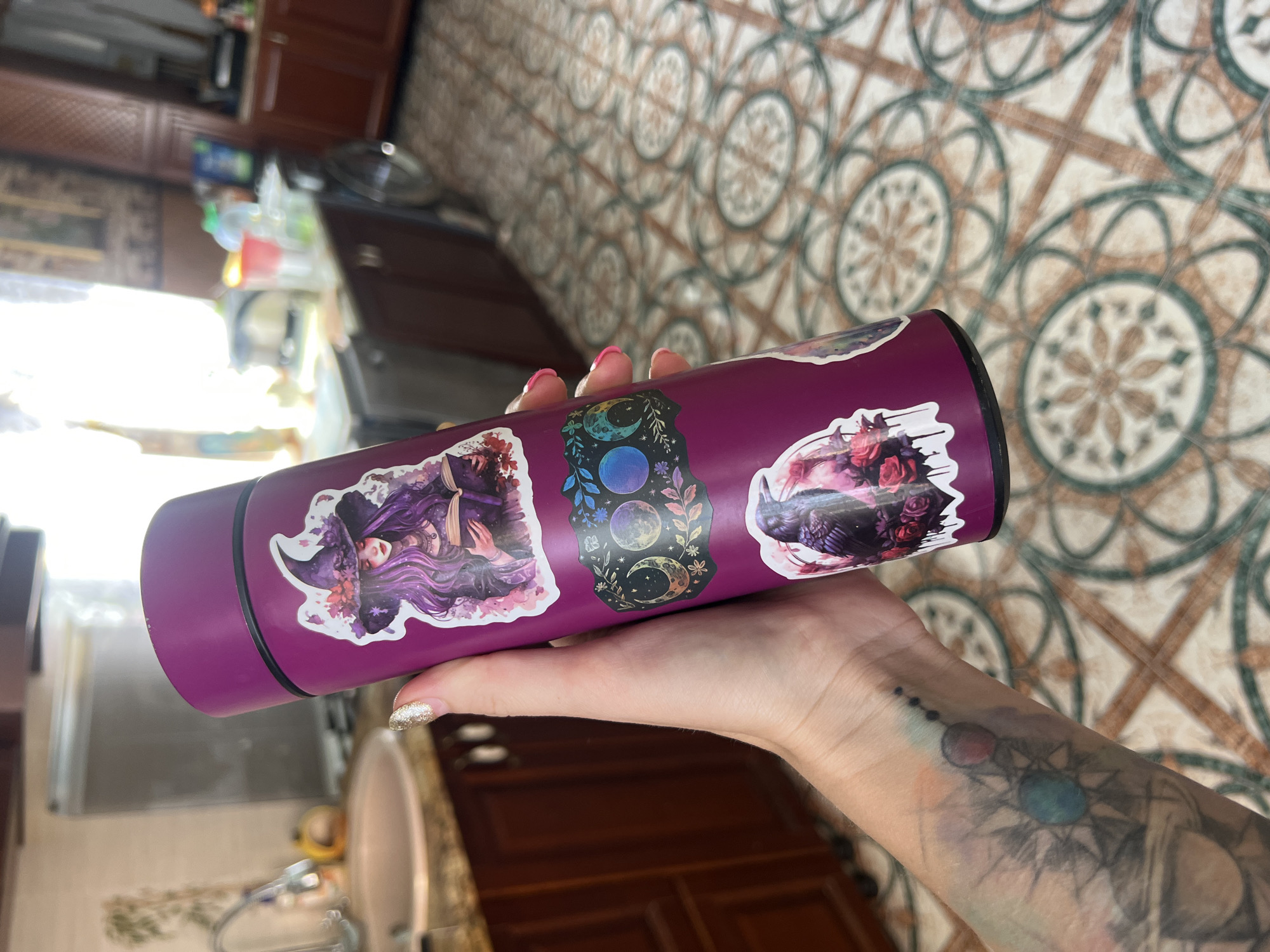 A close-up, top-down view shows a person's hand holding a purple water bottle adorned with several stickers. The stickers feature a witch with purple hair and hat, a celestial design with moons and stars, and a bouquet of pink and red roses with a raven. The person's hand has glittery nail polish and a tattooed forearm with abstract designs, including what appears to be a sun or star motif. The background consists of a patterned tiled floor and a kitchen counter with cabinets visible. The lighting suggests it might be daytime, with light streaming from the left. The scene appears to be indoors, likely in a kitchen or dining area, given the cabinetry and tiled floor. There is no visible text.
