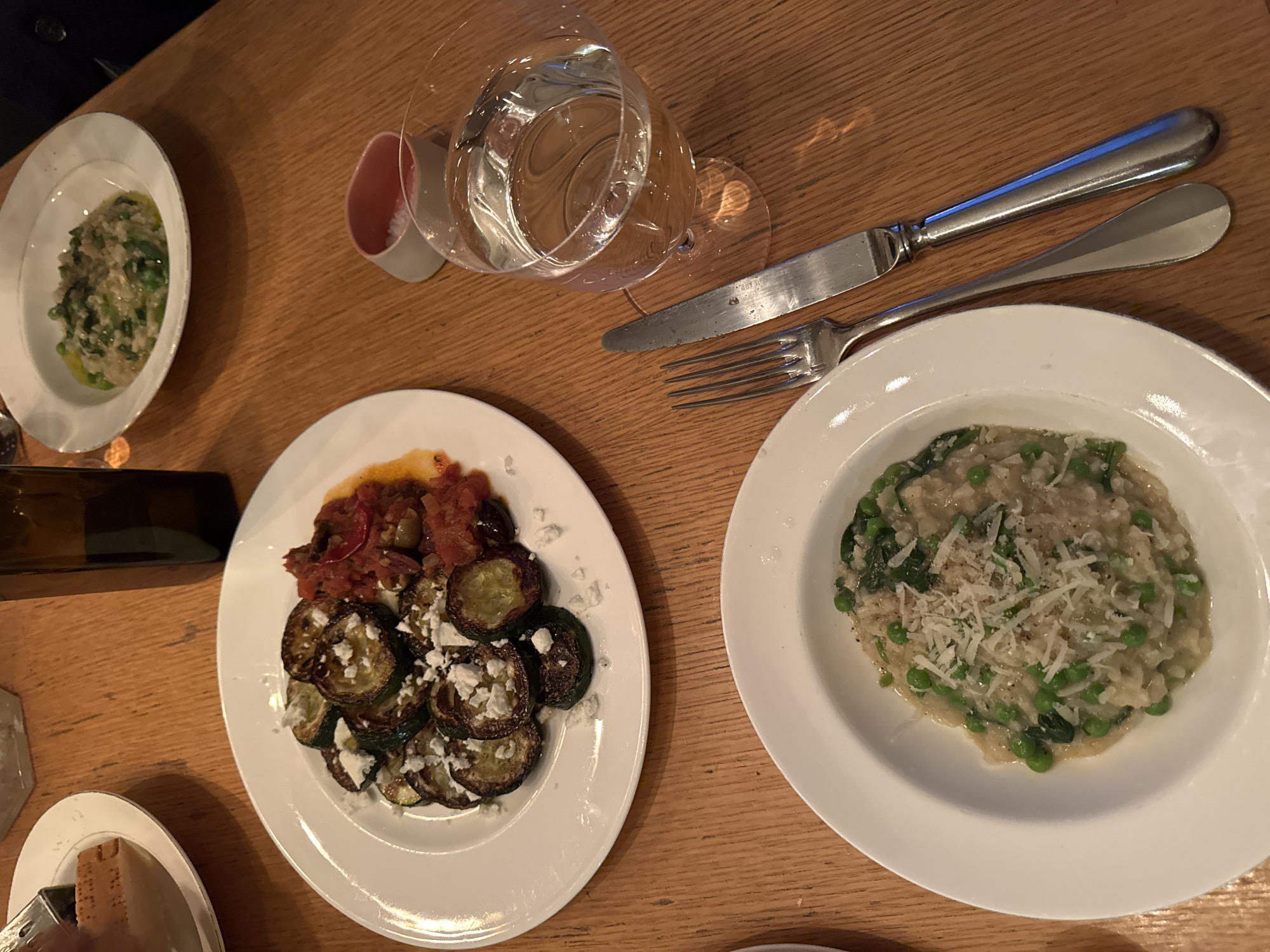 A top-down shot shows a wooden table setting with plates of food, cutlery, and glasses.
Specifically, there are three white plates of food. One plate contains what looks like green risotto with spinach and shaved parmesan cheese. Another has grilled zucchini slices topped with feta cheese and a tomato relish. The third plate, which appears to be a smaller dish, contains a green vegetable dish.
Also visible are a knife and fork, a water glass with water, and a small container filled with a white substance, potentially salt.
The details give the impression of a meal at a restaurant. The location in New York City, United States, is not discernable from the image.