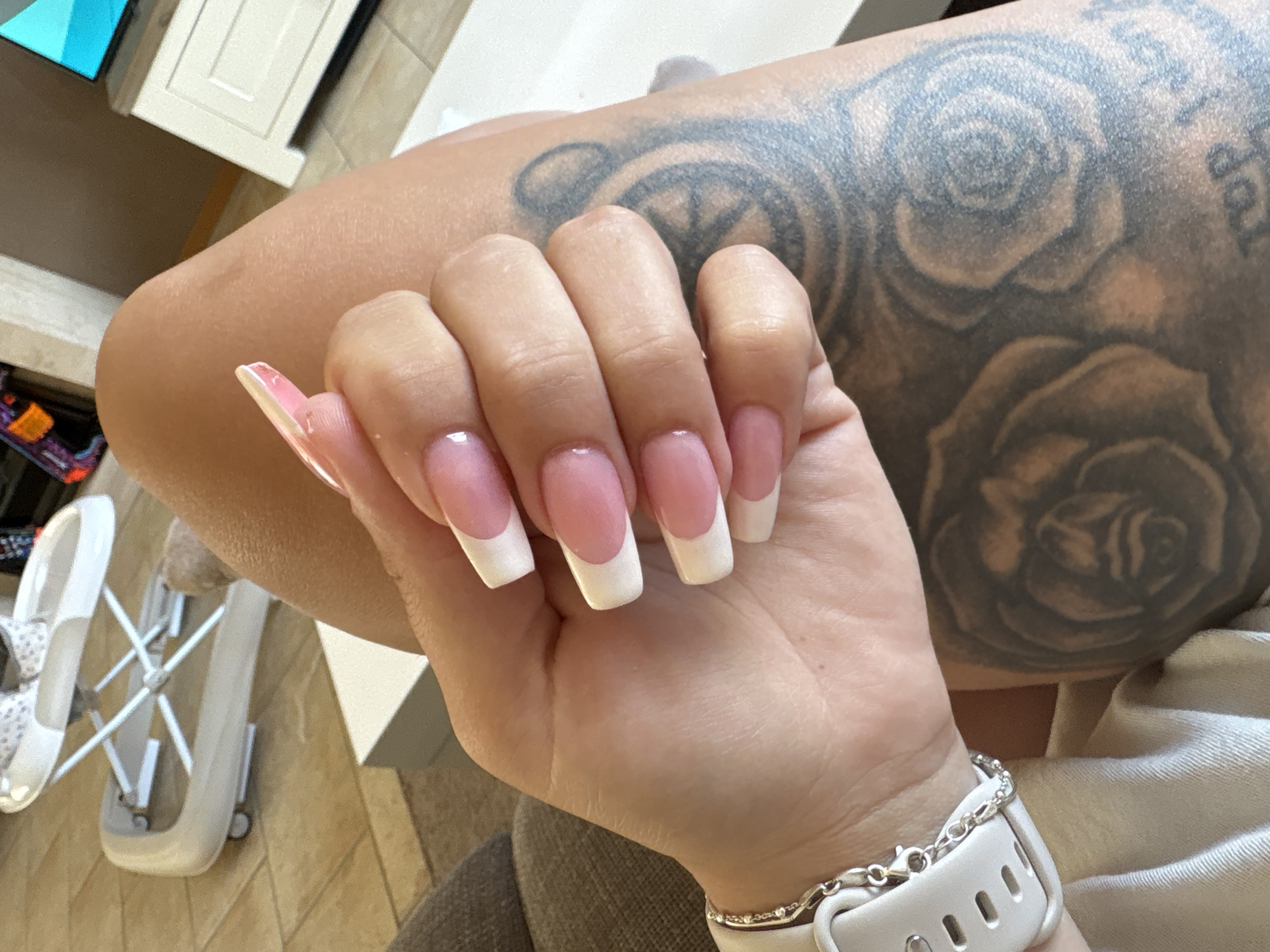 The image shows a close-up of a person's hand with freshly done French tip acrylic nails. The nails are long and square-shaped, with a pink base and white tips. The person's arm, with visible tattoos of roses, is also in the frame. A white smartwatch with a silver chain bracelet is on the person's wrist. In the background, a white baby walker and a glimpse of what appears to be a TV screen and cabinet can be seen. The floor tiles suggest an indoor setting. The lighting indicates it's daytime. No specific emotions are conveyed, and no text is legible in the image. The location is identified as Veghel, Netherlands.