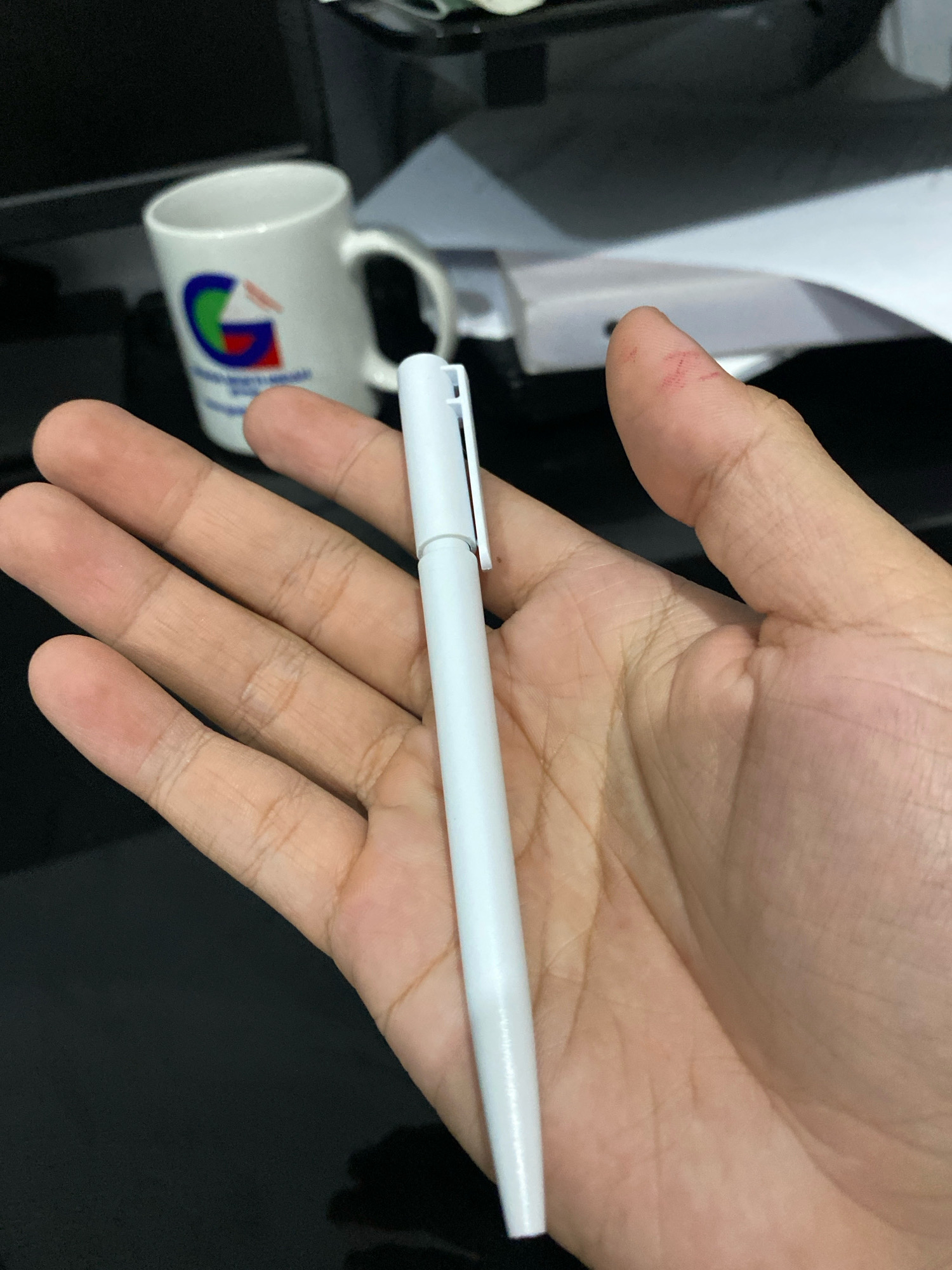 A person's hand is holding a white pen. In the background, a white mug with a logo and some papers are visible. The scene appears to be indoors, possibly at a desk. There are no specific details or objects in the image that indicate a location in Klurak, Indonesia.