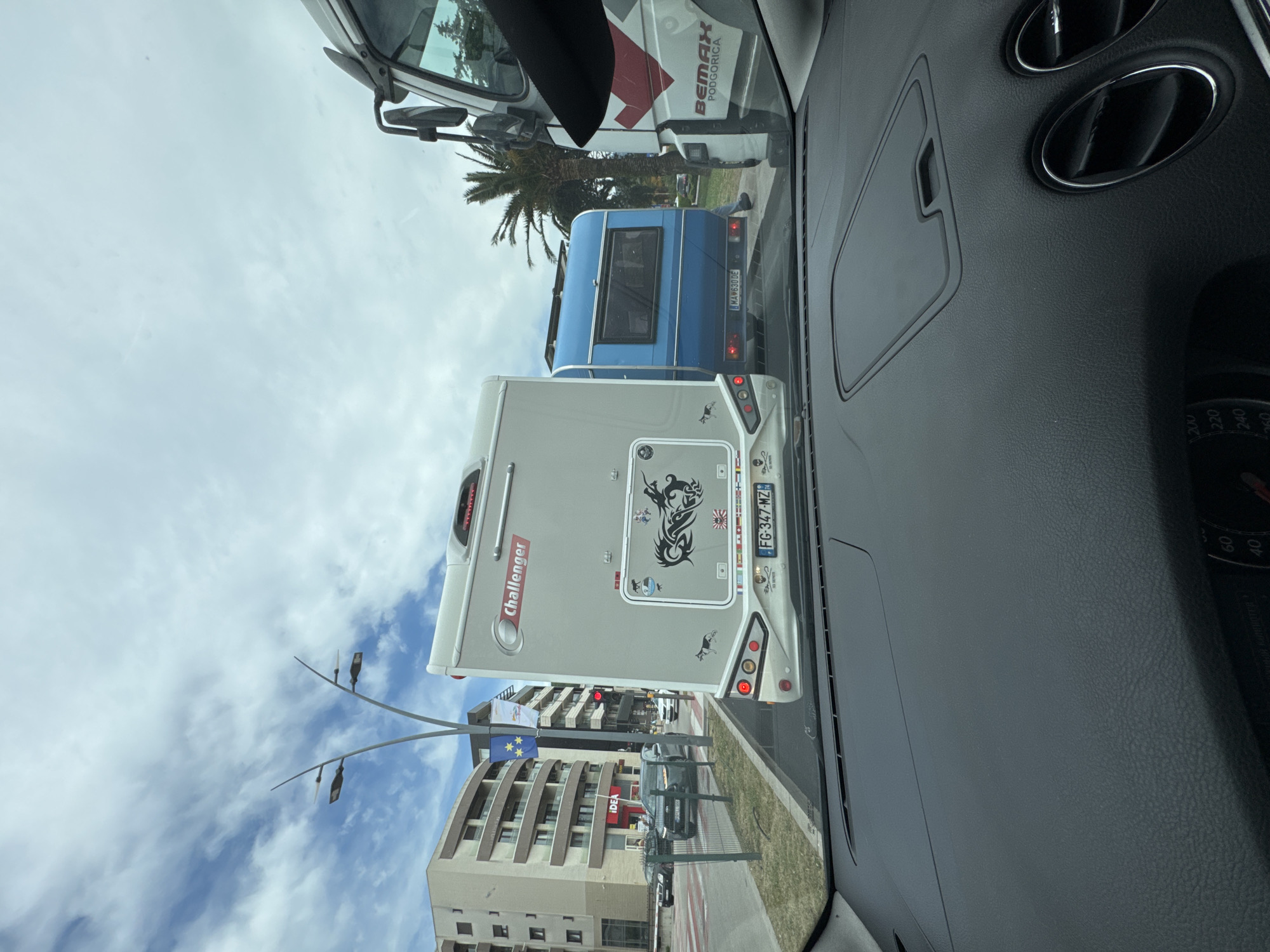 The image, taken from the interior of a car, shows a view through the windshield of the street in Budva, Montenegro. In front of the car is a "Challenger" brand camper with license plate FG-347-MZ. Further ahead is a blue truck with the license plate MA 630 DE and a "BEMAX PODGORICA" marked truck. On the left side of the road, there are buildings, street lights, and the "IDEA" grocery store. European Union and Montenegrin flags are visible. The sky is partly cloudy.