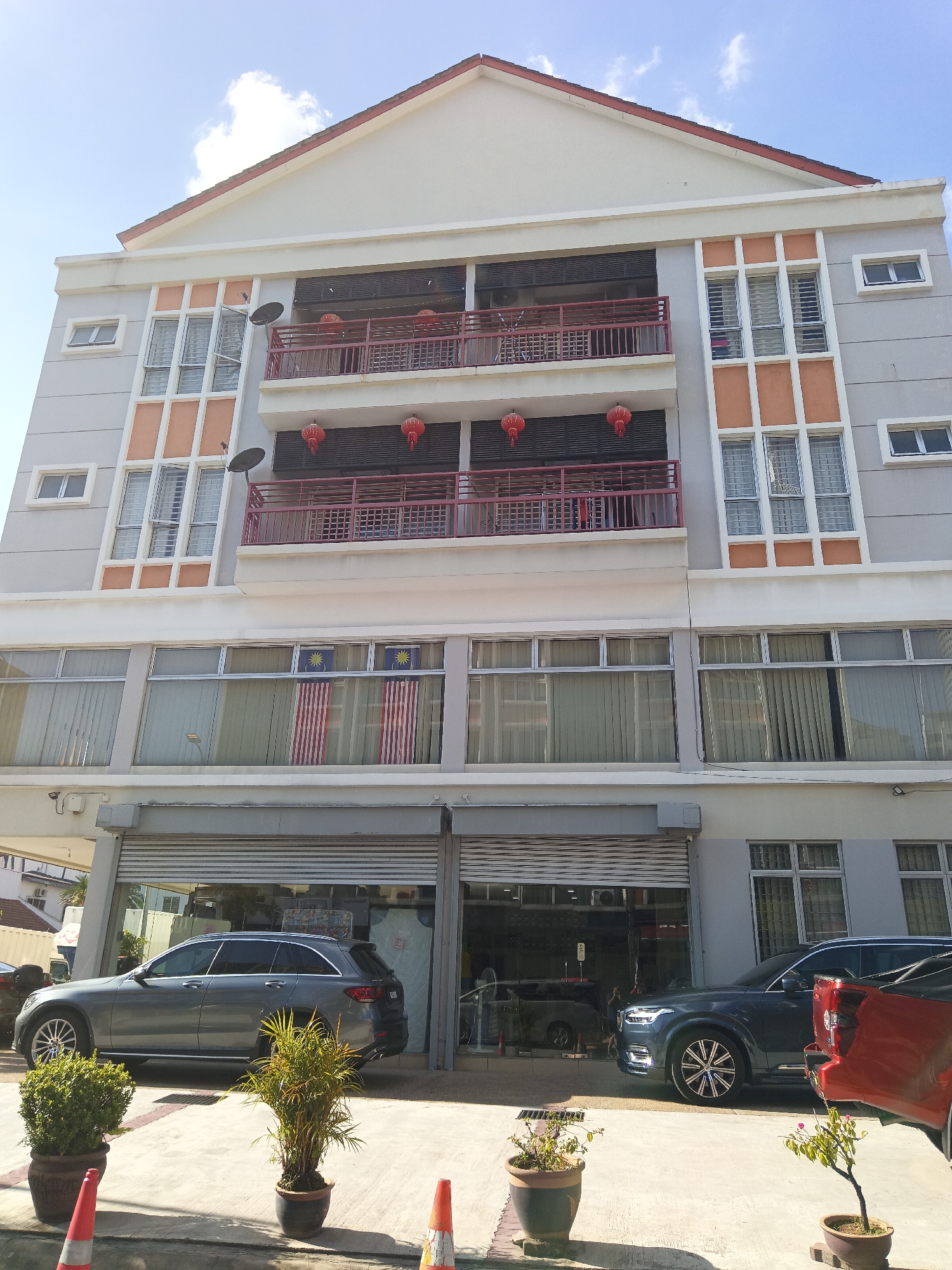 The image shows a multi-story building with a light gray facade and orange accents around some of the windows. The building has multiple levels of balconies with red railings. Red lanterns are hanging from the balconies. The windows on the second floor from the bottom have Malaysian flags displayed in them. On the ground floor, there are shops with roller shutters. Two cars are parked in front of the shops: a dark gray SUV on the left and a dark blue SUV on the right. Potted plants are placed on the pavement in front of the building. Orange traffic cones are also visible on the pavement. The sky is blue with some white clouds. The scene appears to be in a commercial area of Kuala Lumpur, Malaysia, given the presence of the Malaysian flags.