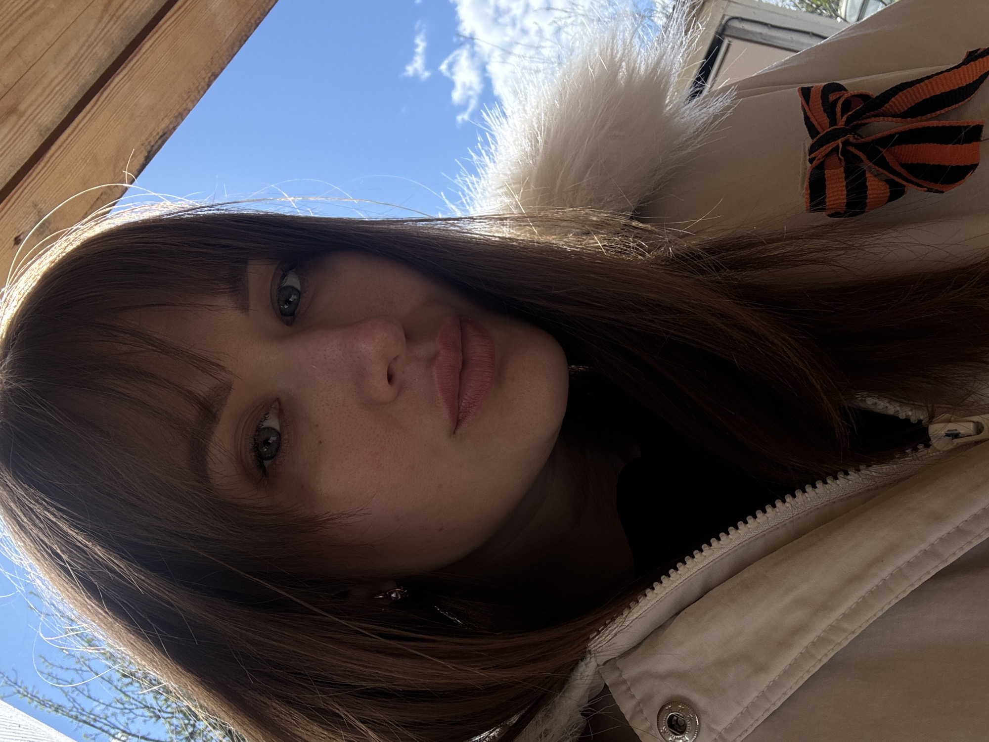 A medium shot depicts a woman with light skin, brown hair with bangs, and green eyes. She is wearing a beige jacket with a fur-trimmed hood and a black-and-orange ribbon pinned to the jacket. The photo is taken from a slightly low angle, looking up at the woman. The background shows a blue sky with some clouds and trees. There is no information available to confirm the location as the city of Chaikovsky, Russia.