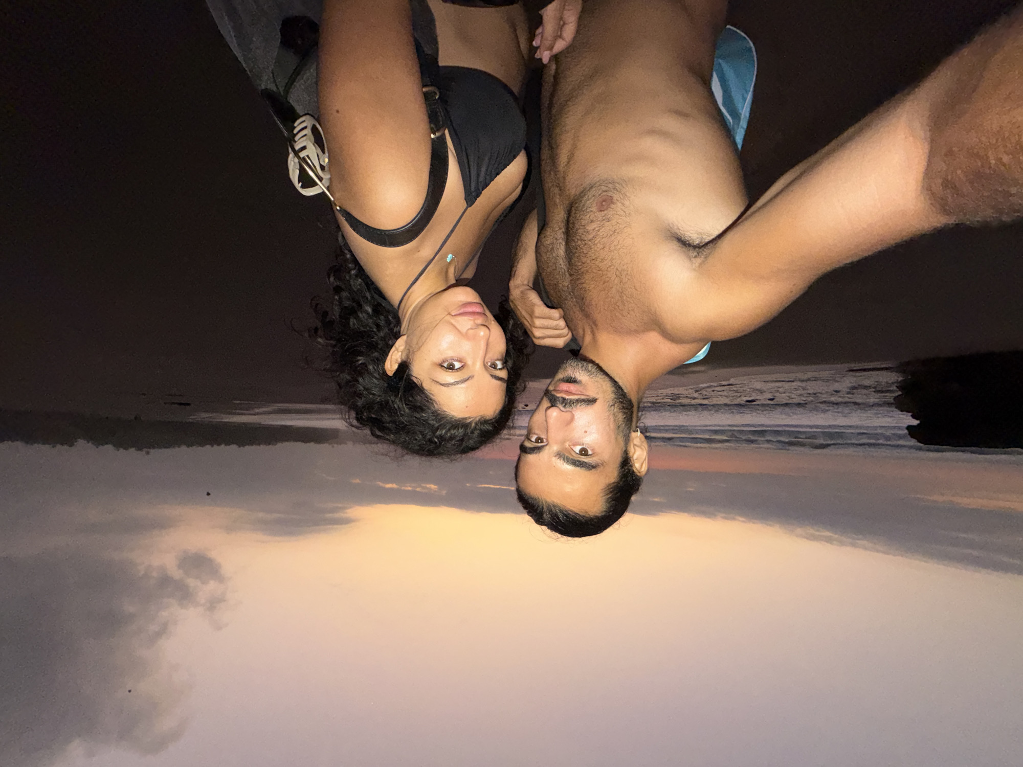 This image captures two individuals, a male and a female, posing for a selfie on a beach in Selemadeg Timur, Indonesia, during sunset or dusk.
The male subject, positioned on the left, has dark skin, dark hair pulled back, a beard, and a mustache. He wears a small silver hoop earring in his left ear and is bare-chested, revealing chest hair and a lean physique with visible ribs. A light blue towel or fabric is draped over his left shoulder and arm. His right arm is extended, holding the camera. His left hand is resting on or near the female subject's waist, partially obscured. He maintains direct eye contact with the camera.
The female subject, on the right, has dark skin, dark curly hair, and wears a black bikini top. A thin necklace with a small turquoise pendant is visible around her neck. A black strap, likely from a bag or backpack, is slung over her right shoulder, with a light-colored, possibly decorative, object attached to the strap on her back. Her left hand is positioned near the male subject's waist, partially obscured by his arm. She also looks directly at the camera with a slight, subtle expression.
The environment depicts a wide sandy beach. The ocean is visible to the left, with waves breaking on the shore. A dark, rocky outcrop is present on the far left of the beach. Distant land with dark vegetation is discernible on the far right, across the beach. The sky exhibits the colors of sunset or dusk, with warm orange and pink tones near the horizon gradually fading into cooler purples and greys overhead. Large, elongated dark grey clouds dominate much of the sky. Two small, dark, indistinct flying objects, possibly birds or kites, are visible against the clouds in the upper right quadrant of the image.