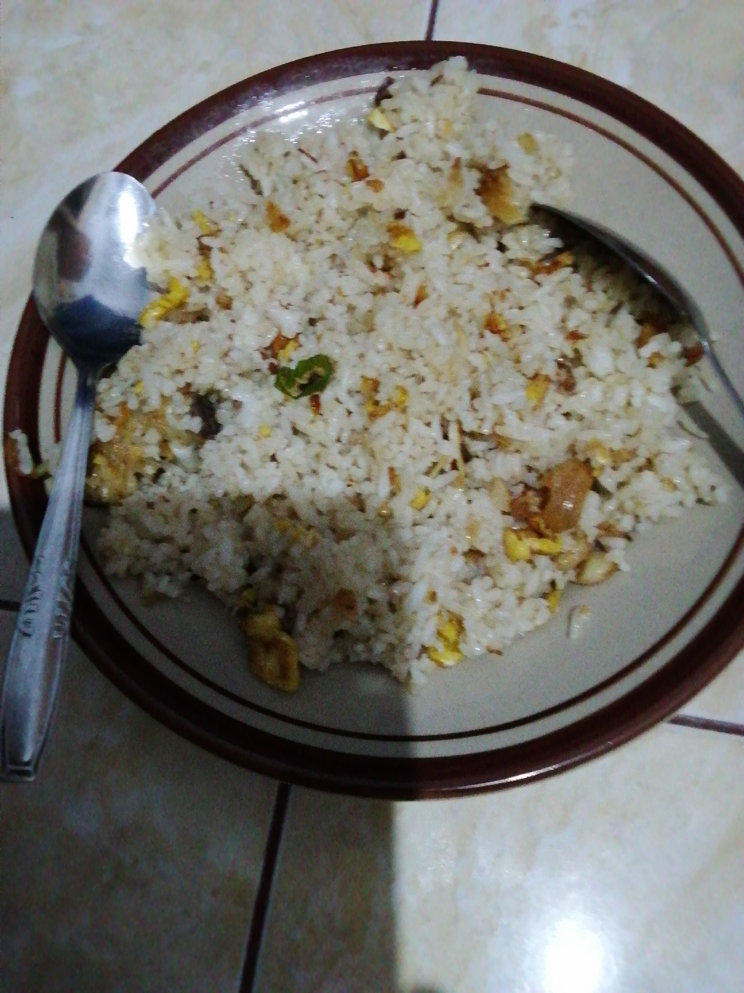 The image shows a close-up view of a plate of fried rice served on a patterned tile surface. Two spoons are visible, one on the left side of the plate and another partially submerged in the rice on the right. The fried rice appears to be a simple preparation with visible grains of rice, scrambled egg pieces, and possibly small bits of meat or vegetables. The plate has a dark rim with a lighter stripe. The lighting suggests an indoor setting, possibly a home or a casual dining establishment. There are no people visible in the frame. The overall scene depicts a meal ready to be eaten. The floor surface is made of light-colored tiles with darker grout lines. The image is taken from a slightly elevated angle, looking down onto the plate of food.