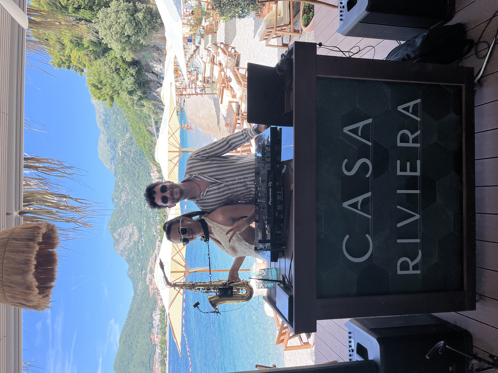 The image depicts a DJ booth and two individuals on a wooden deck at the "Casa Riviera" establishment, situated on the beach in Budva, Montenegro. The beach features numerous sun loungers and umbrellas, and the turquoise sea stretches out towards a backdrop of lush green mountains.
In the foreground, a DJ booth displays a Pioneer DJ controller and a laptop. To the left of the booth, a woman is holding a saxophone, wearing sunglasses and a white top. To her right, a man in a striped shirt and sunglasses stands near the DJ equipment.
Behind the DJ booth, the beach is visible with people relaxing on loungers and swimming in the sea. The clear blue sky suggests a sunny day, and the overall atmosphere appears to be one of leisure and entertainment. The "Casa Riviera" logo is prominently displayed on the front of the DJ booth. The presence of sound equipment, including speakers on either side of the booth, indicates that music is being played or is about to be played. The natural beauty of the mountainous coastline and the beach setting contribute to the idyllic scene.
