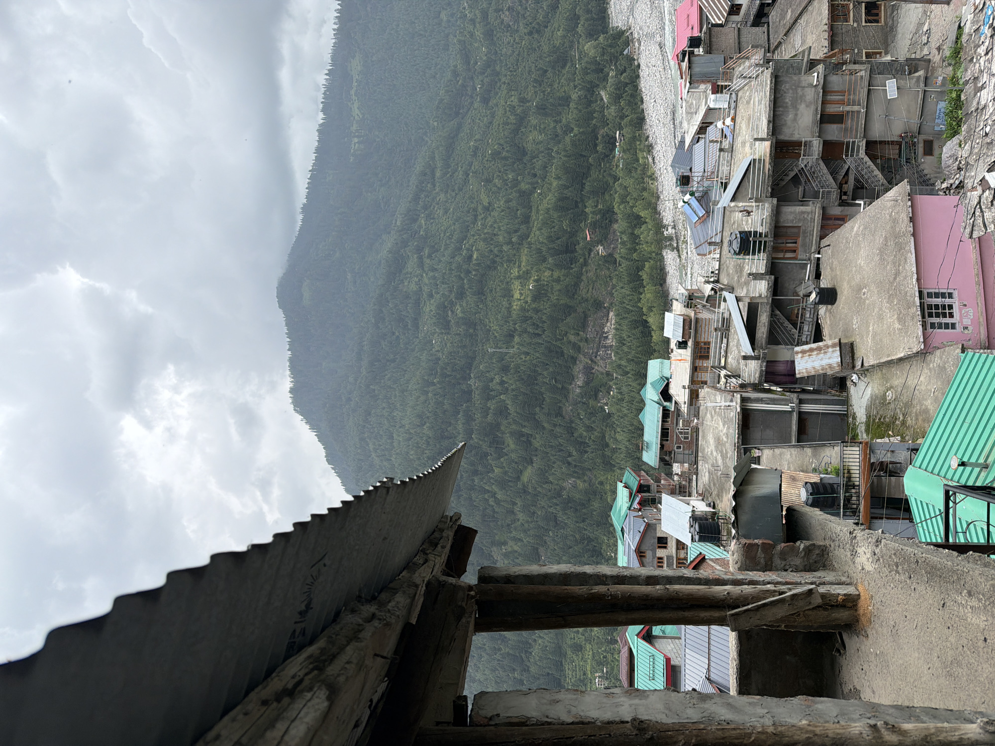 This is an outdoor, medium-distance, eye-level shot taken in Vashisht, India, under an overcast sky. In the foreground, a partially constructed building with rough wooden beams and a corrugated metal roof is visible on the left. Beyond this structure, a cluster of multi-story buildings with concrete facades and a mix of green, pink, and gray roofs fills the midground. Some of these buildings have external staircases and water tanks on their rooftops. In the background, a dense, forested mountainside slopes upwards, with a riverbed visible at the base of the mountain. The overall scene depicts a village nestled in a mountainous region.