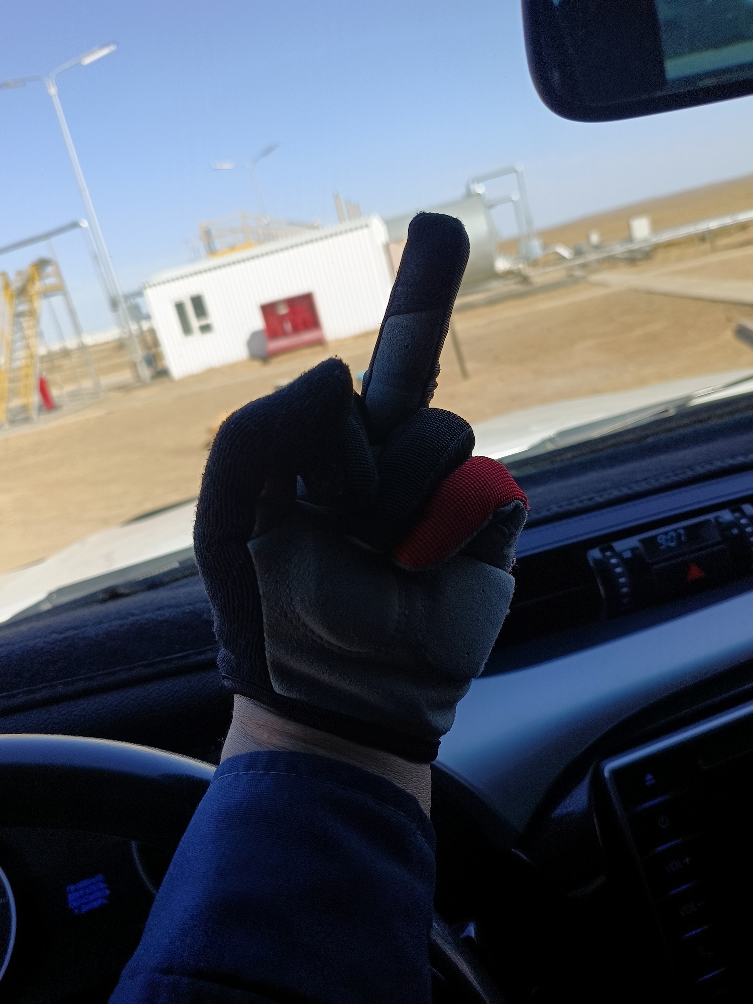 A gloved hand is raised, middle finger extended, in the foreground of the image. The hand wears a dark blue glove with grey and red accents on the fingers. The background shows an industrial site with a white building, a large silver tank, and yellow scaffolding under a clear blue sky. The photo is taken from inside a vehicle, with part of the dashboard and steering wheel visible in the lower portion of the frame. The time on the car's display reads 9:07.