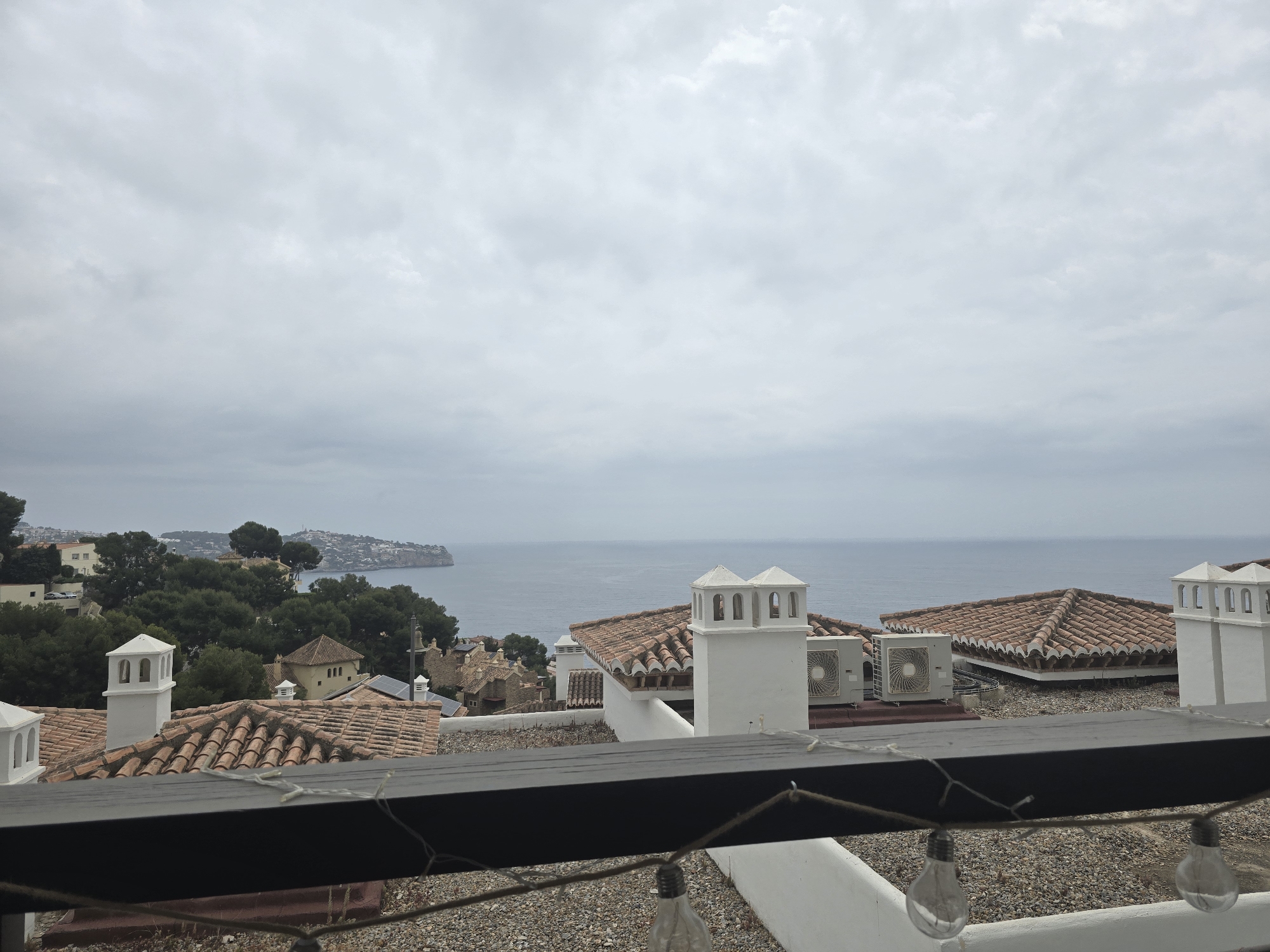 The image shows a coastal scene in Almuñécar, Spain, taken from an elevated vantage point. A balcony with a dark-colored railing and hanging light bulbs frames the view. In the middle ground, there are rooftops with terracotta tiles and white chimneys. Some air conditioning units are visible on the rooftops. Beyond the rooftops, the sea stretches out to the horizon under a cloudy sky. A landmass with vegetation is visible in the distance to the left.