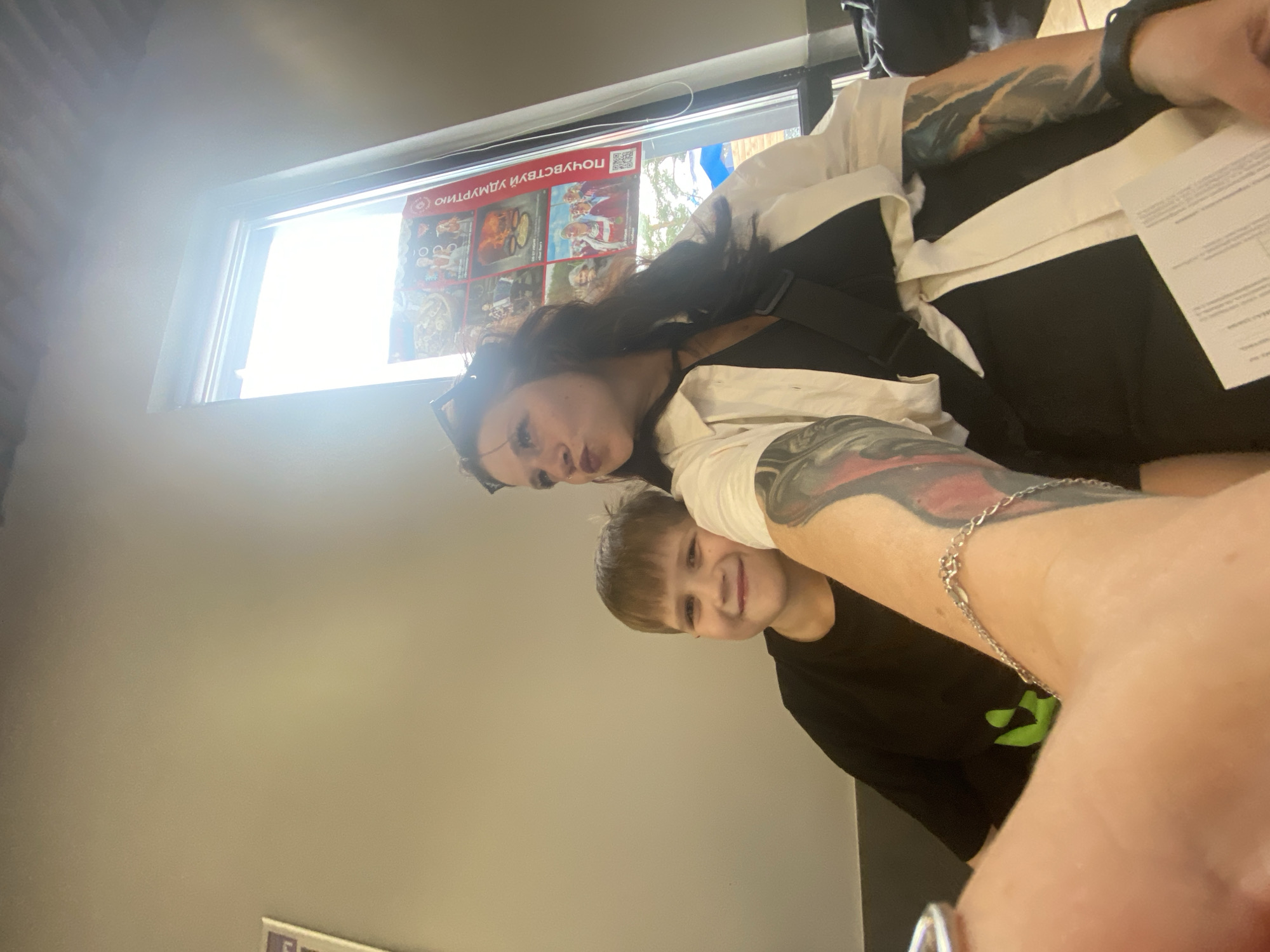 In this selfie, a woman with dark hair and tattoos on her arm is taking a photo with a young boy. The woman is wearing a white top over a black garment. The boy, with short brown hair, is smiling and looking at the camera. He is wearing a black t-shirt with a green logo. The scene appears to be indoors, possibly a store or public space, with a large window in the background. The window shows a bright, sunny day with some trees visible outside. On the window, there are posters with images and text. One poster displays a QR code and text that appears to be in Cyrillic. The overall mood is casual and happy, with both individuals smiling for the photo. The image is taken from a slightly tilted angle, typical of a selfie.