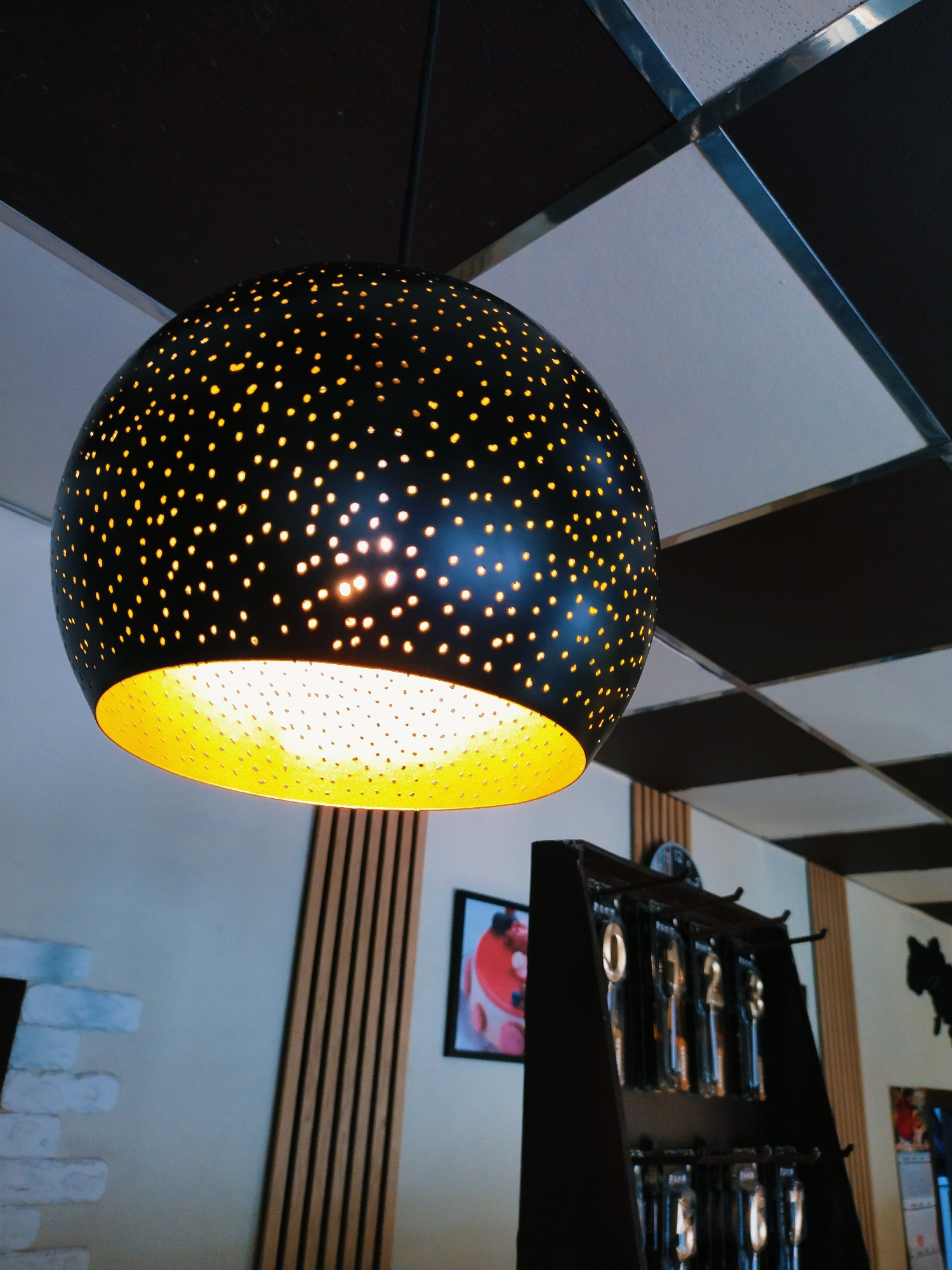 This media file shows a close-up view of a black, spherical pendant light with a perforated design, allowing a warm yellow light to shine through. The light fixture is hanging from a dark ceiling composed of square panels, alternating between black and white. The background reveals a wall with a decorative element of vertical wooden slats, and further to the right, a dark wooden display stand holding several items in clear packaging, possibly cutlery or tools, each with a label. There is also a framed picture on the wall depicting a cake with fruit. The overall scene suggests an indoor setting, possibly a restaurant, cafe, or retail space. The lighting and ambiance suggest it could be evening or a dimly lit interior. No people are visible. There is no discernible weather or time of day information. Location cues for Horishni Plavni, Ukraine are provided but not visually evident in the image itself.