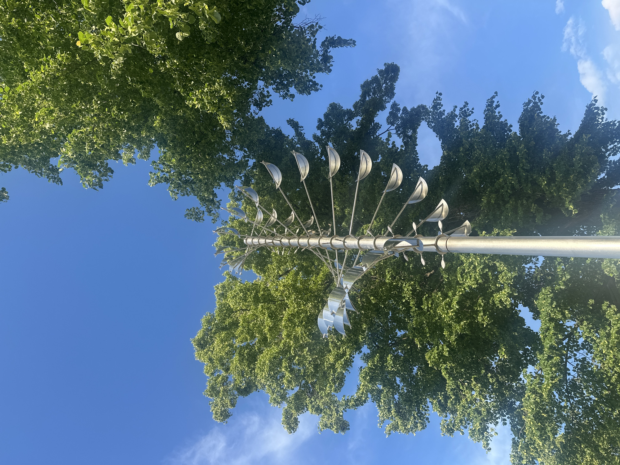 The image shows a low-angle view of a metal pole-like structure with multiple wind-catching elements resembling stylized leaves. The structure is positioned between two rows of trees with dense green foliage, set against a bright blue sky with some wispy clouds. The angle of the shot suggests the viewer is looking upwards from the ground.
Based on the description, it is likely the object is a decorative wind sculpture commonly seen in urban environments such as Votkinsk, Russia.