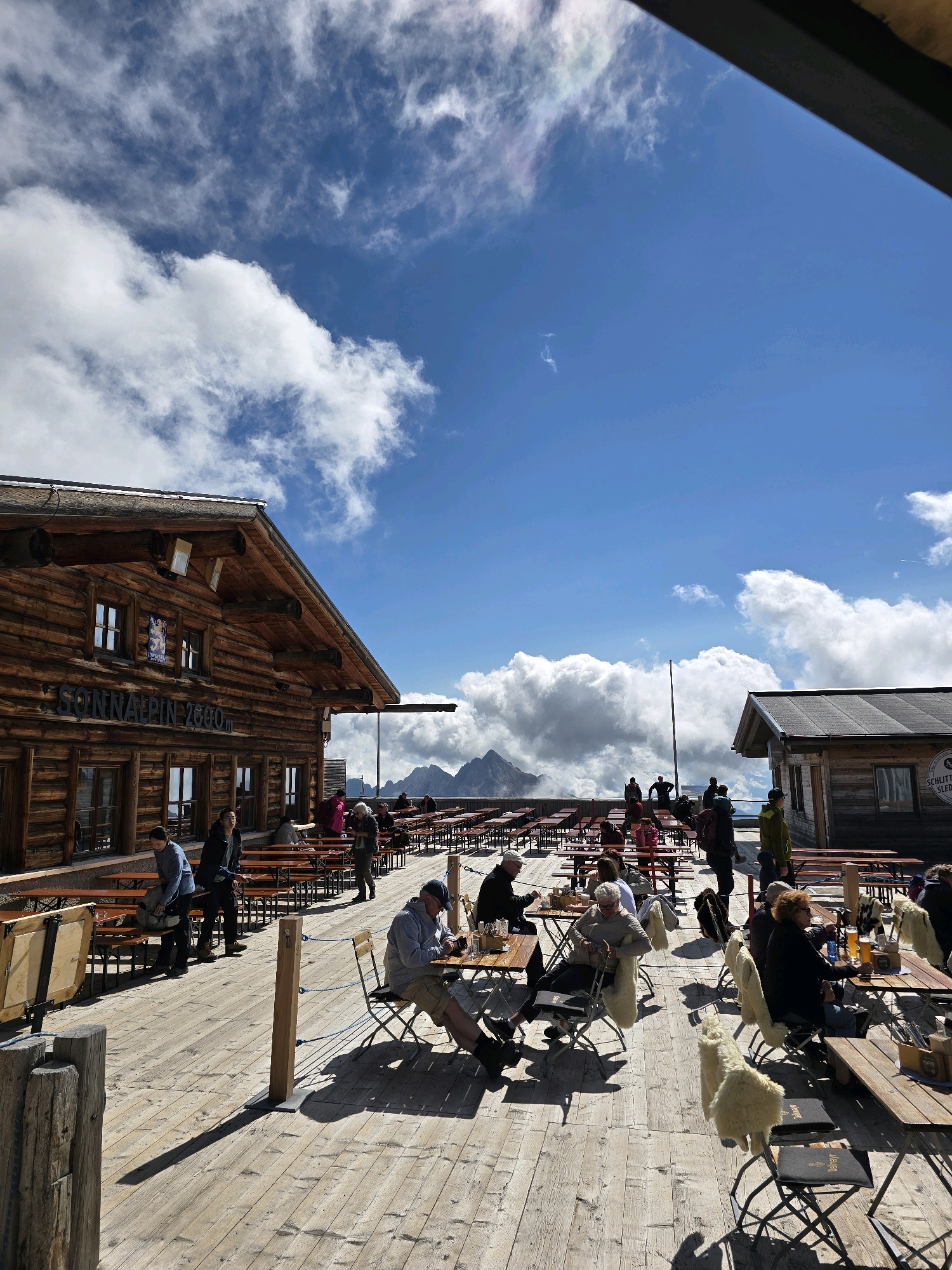The image depicts an outdoor seating area of a mountain restaurant, likely at the "SonnAlpin 2000m" location, situated in the Alps near Garmisch-Partenkirchen, Germany. The scene is dominated by a large wooden building with several windows and the text "SONNALPIN 2000m" visible on its facade. Numerous wooden tables and benches are set up on a wooden deck, with people seated at some of them, enjoying drinks and presumably a meal.
The sky is a vibrant blue, with scattered white clouds. In the background, mountains are visible through a gap in the clouds. The lighting suggests it is daytime, with the sun casting shadows on the wooden deck.
People are casually dressed, indicative of a tourist destination. Some are seated, engaged in conversation or looking at their phones, while others are standing or walking around. The atmosphere appears relaxed and social. The wooden structures and the mountain landscape contribute to a rustic, alpine setting.