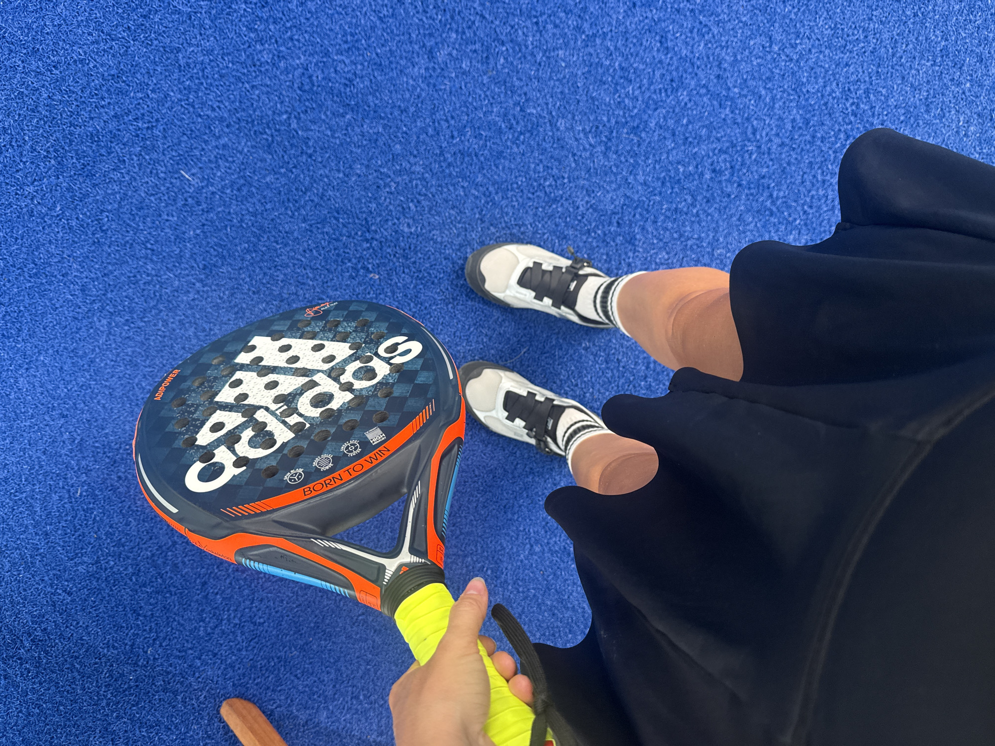 A person's legs and torso are visible, wearing a black skirt, white socks with black stripes, and white and gray athletic shoes. They are standing on a blue artificial turf surface. The person is holding a dark blue and orange padel racket with a bright yellow grip. The racket has the "adidas" logo and the text "BORN TO WIN" printed on it. A small portion of a wooden chair is visible in the lower right corner. The scene appears to be an outdoor sports court, possibly in the city of Ban Bang Rak, Thailand, given the context.