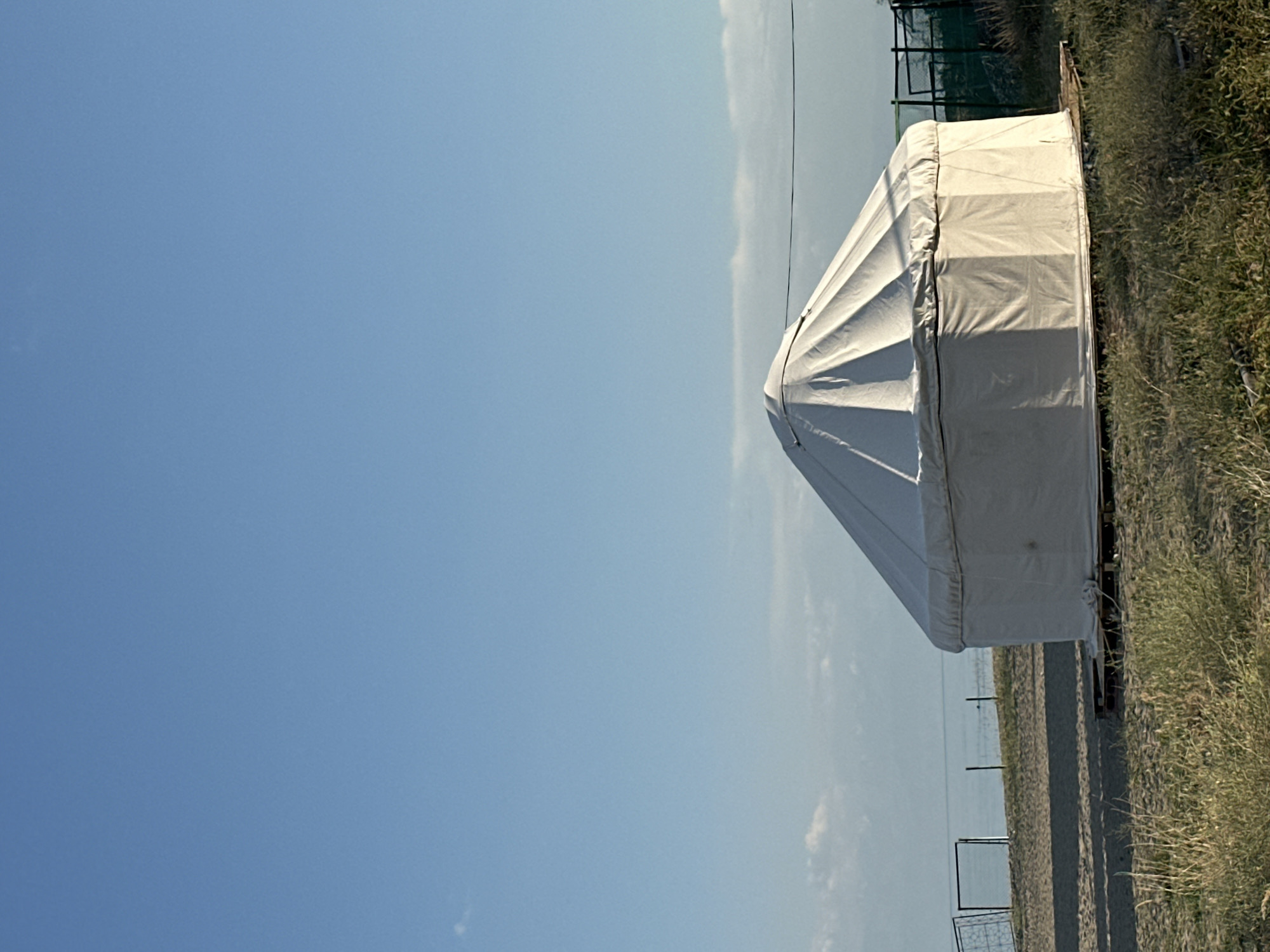 The image shows a white yurt situated on a sandy shore in Grigorevka, Kyrgyzstan. The yurt is positioned on wooden pallets, with the surrounding area covered in dry grass and sand. In the background, a body of water, likely Issyk-Kul Lake, is visible, with a clear blue sky above. There are also fences visible along the shoreline, suggesting a boundary or enclosure. The lighting indicates it is daytime, with the sun casting shadows. No people or animals are present in the frame.