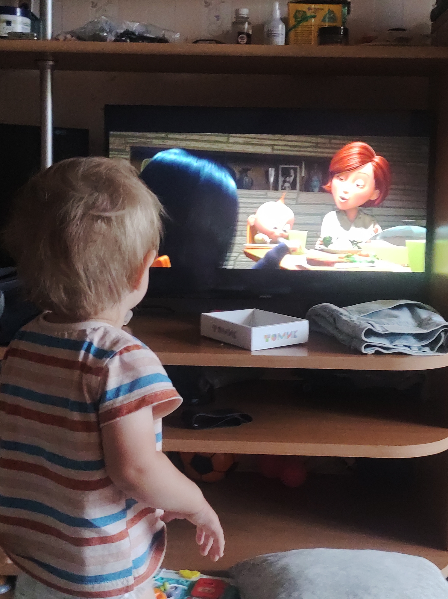 A toddler with blond hair is watching a television. The screen shows an animated scene with characters from "The Incredibles." The toddler wears a striped shirt. The television sits on a shelf with a white box and a pair of gray pants. There are other items on the shelf above. The location is Chaikovsky, Russia.