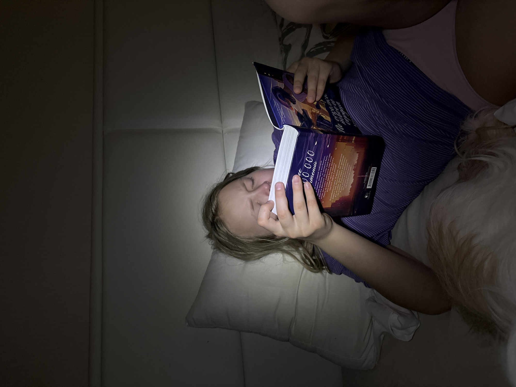 A girl lies on a white couch, reading a book tilted in her hands. The book cover is titled "10,000..." in what appears to be Cyrillic script. The girl has blond hair and is wearing a purple striped shirt and pink underwear. The location, Izhevsk, Russia, cannot be confirmed from the given image.