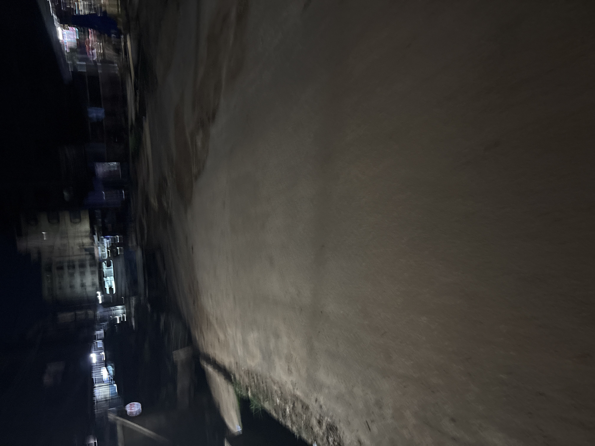 The image captures a blurry, nighttime view of a street in Onitsha, Nigeria. A paved road occupies the majority of the frame, with a dirt and grass embankment visible on the left side. In the background, indistinct buildings and lights suggest an urban environment. A circular red and white sign is visible on the left, near the embankment. The overall impression is one of motion blur, indicating movement during the photograph's capture.