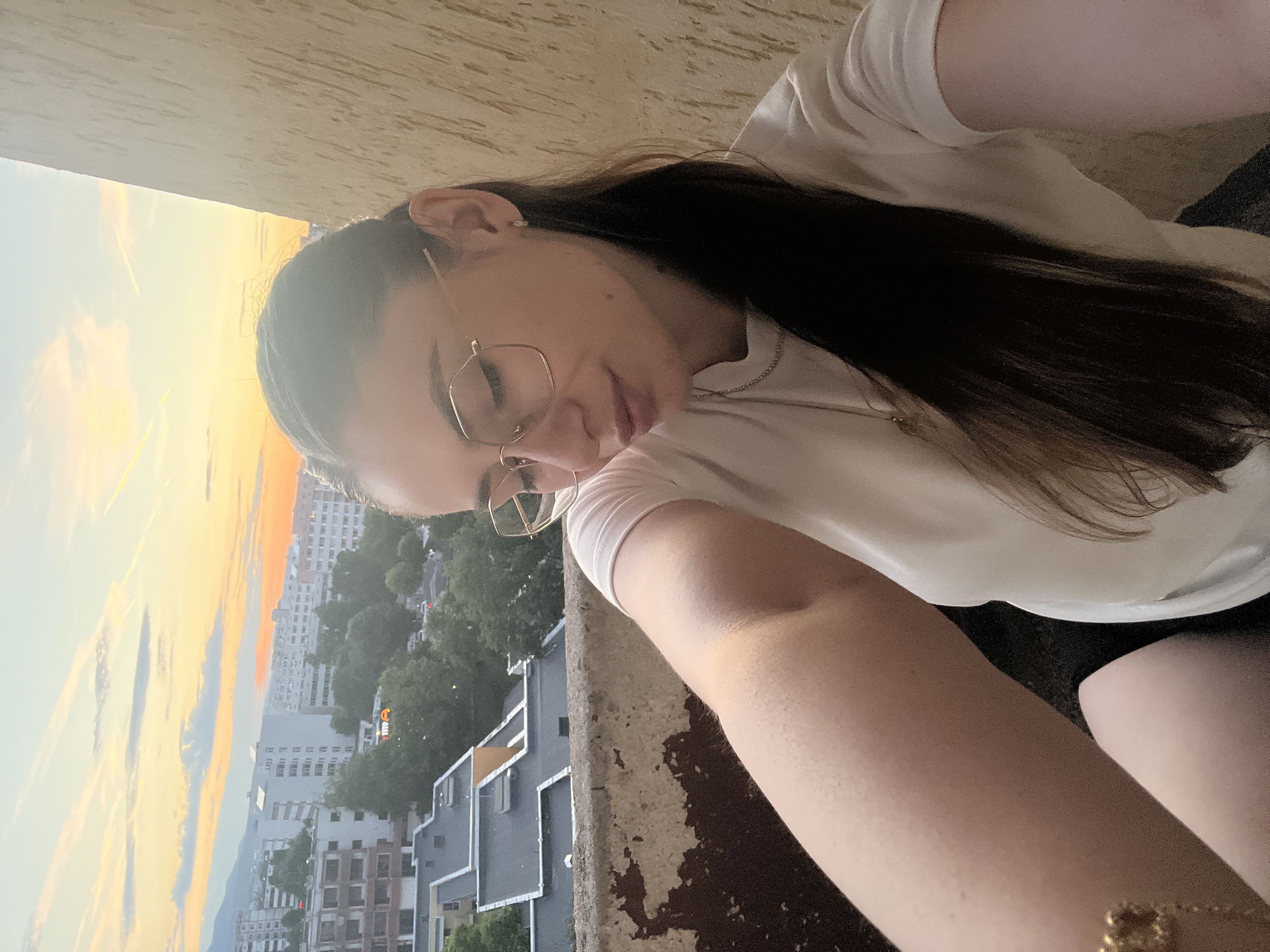 A young woman with long dark hair and glasses takes a selfie on a balcony with a city view at sunset. She is wearing a white t-shirt and a gold bracelet. The buildings and trees of the city are visible in the background, bathed in the warm light of the setting sun. The balcony railing is weathered and cracked.