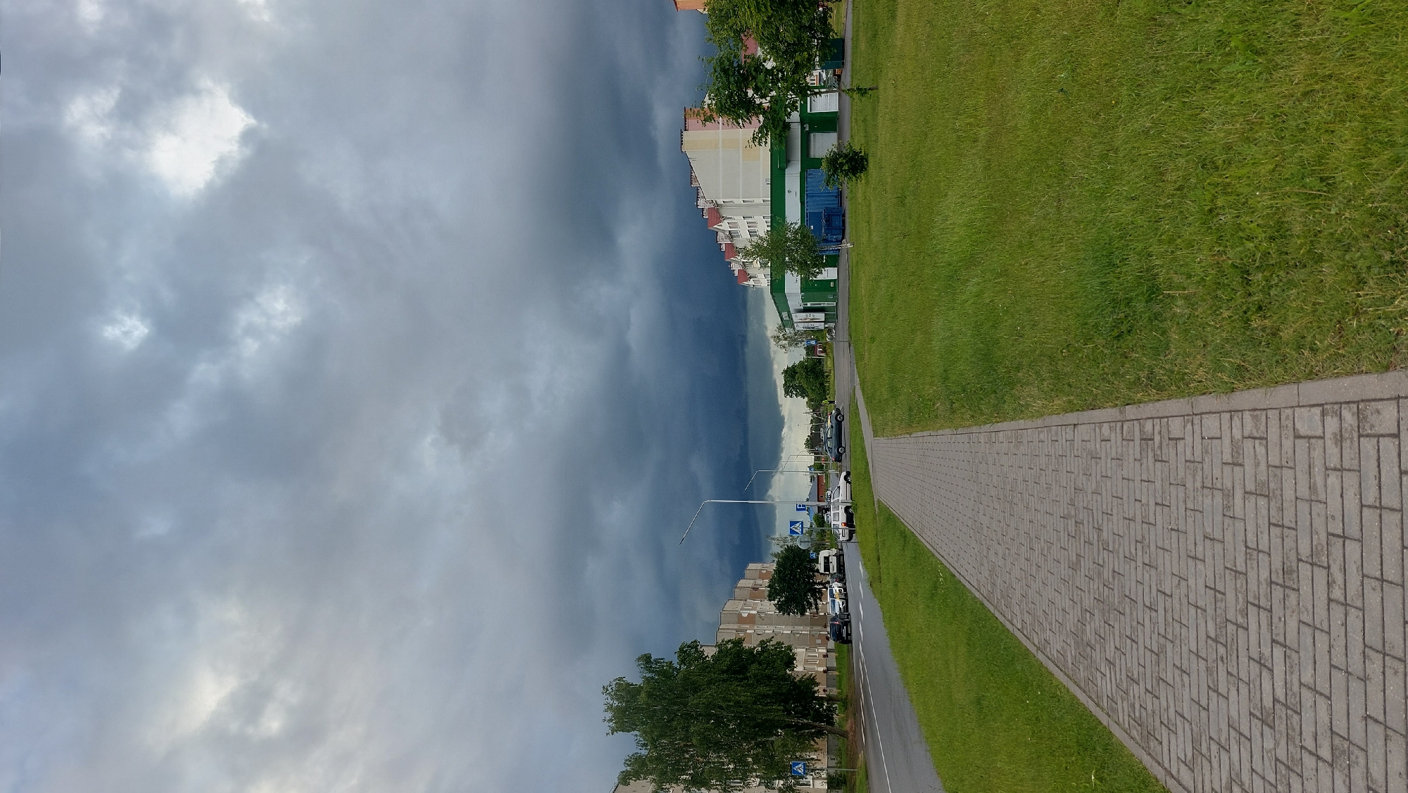The image captures a scene in the city of Masty, Belarus. A paved pedestrian walkway runs along the right side of a road, bordered by a neatly mown grassy area. On the left, a multi-story apartment building lines the road, with a tall green tree in front of it. Further down the road, several cars are parked. On the right side of the road, across from the apartment building, is a low-rise building with a green facade, possibly a store or commercial establishment. Beyond this building, more apartment buildings are visible. The sky is filled with dark, heavy storm clouds, creating a dramatic and overcast atmosphere.