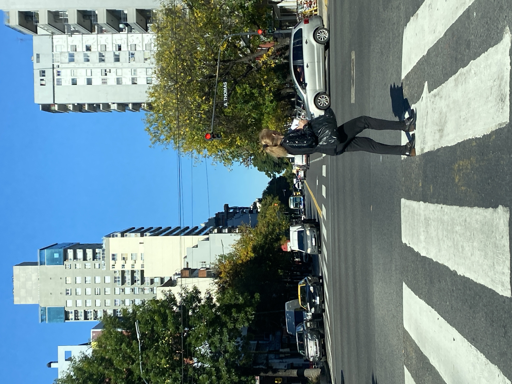 A woman is seen crossing a street in Buenos Aires, Argentina. She has blonde hair and is wearing a black leather jacket and black pants. She is walking on a pedestrian crosswalk. There is a sign on the street that reads "A. Thomas". There are trees and buildings on both sides of the street. The sky is blue. A red traffic light is visible. Cars are parked along the street.