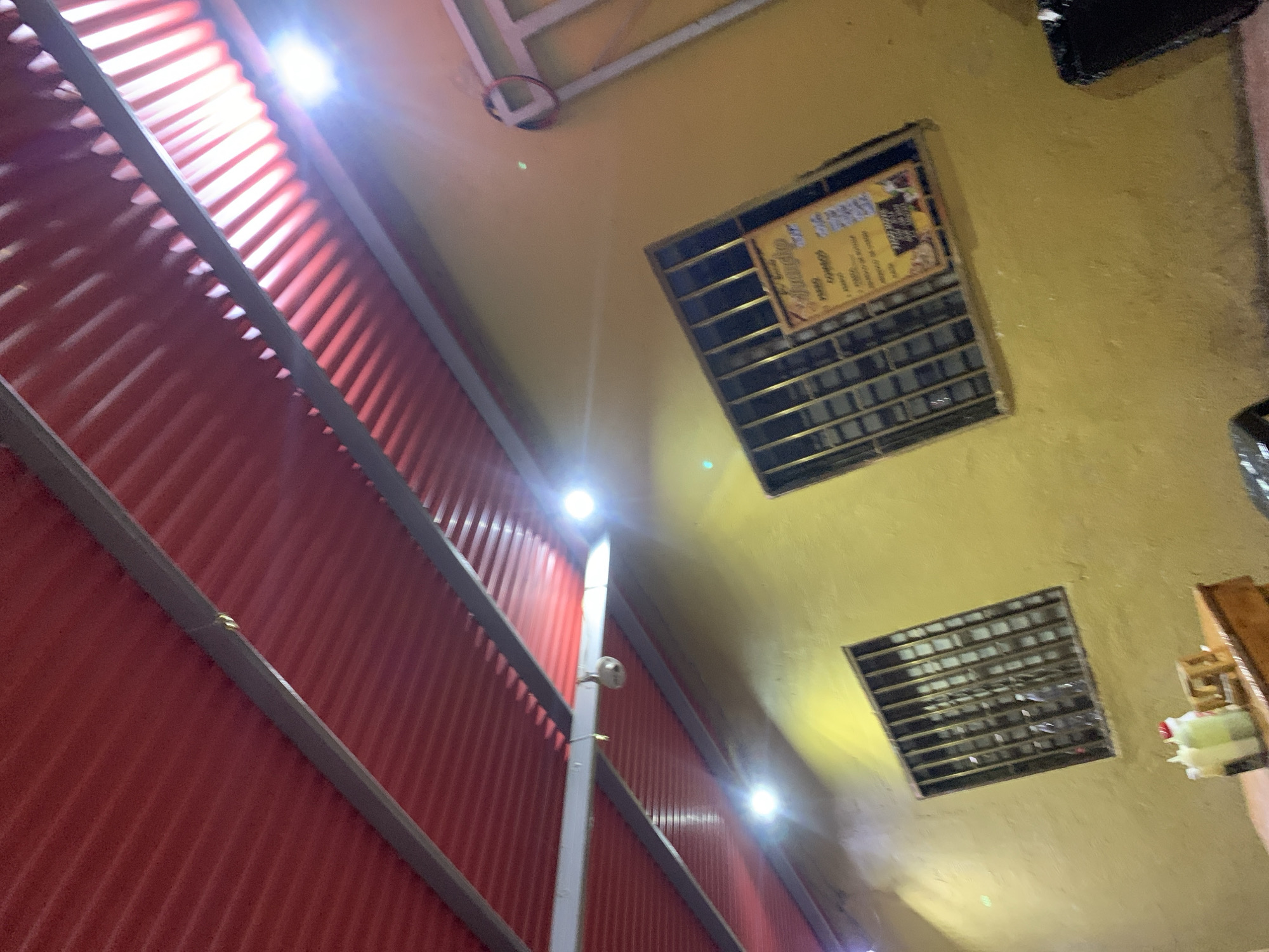 The image captures an upward-looking view inside what appears to be a commercial or public establishment in Santa Ana, Venezuela, likely at night or in an artificially lit setting due to the prominent bright lights.
On the left, a textured red corrugated metal wall stretches vertically, intersected by several horizontal grey metal beams. Along this wall and on a central vertical pole, multiple bright, circular white LED lights are mounted, emitting strong light and producing lens flares. A white CCTV camera is also visible on the central pole, pointed away from the camera.
The ceiling or upper wall area, dominating the right and top portions of the image, is a textured yellow-gold color. Embedded within this surface are two rectangular metal grates or vents. Above the upper vent, a framed sign or poster is partially visible, obscured by a metal grid. Though somewhat unclear, legible text on the sign includes "Chupos" (possibly a name or product), and words like "cafe," "gratis" (free), "fruta," and "jugos" (juices), suggesting it functions as a menu or advertisement for food and beverages.
In the bottom right corner, a glimpse of a wooden structure and some small, colorful objects (possibly bottles or containers) are visible. No people are present, and no specific activity is depicted, lending to a static view of the interior architecture and fixtures. The overall aesthetic is functional and somewhat industrial.