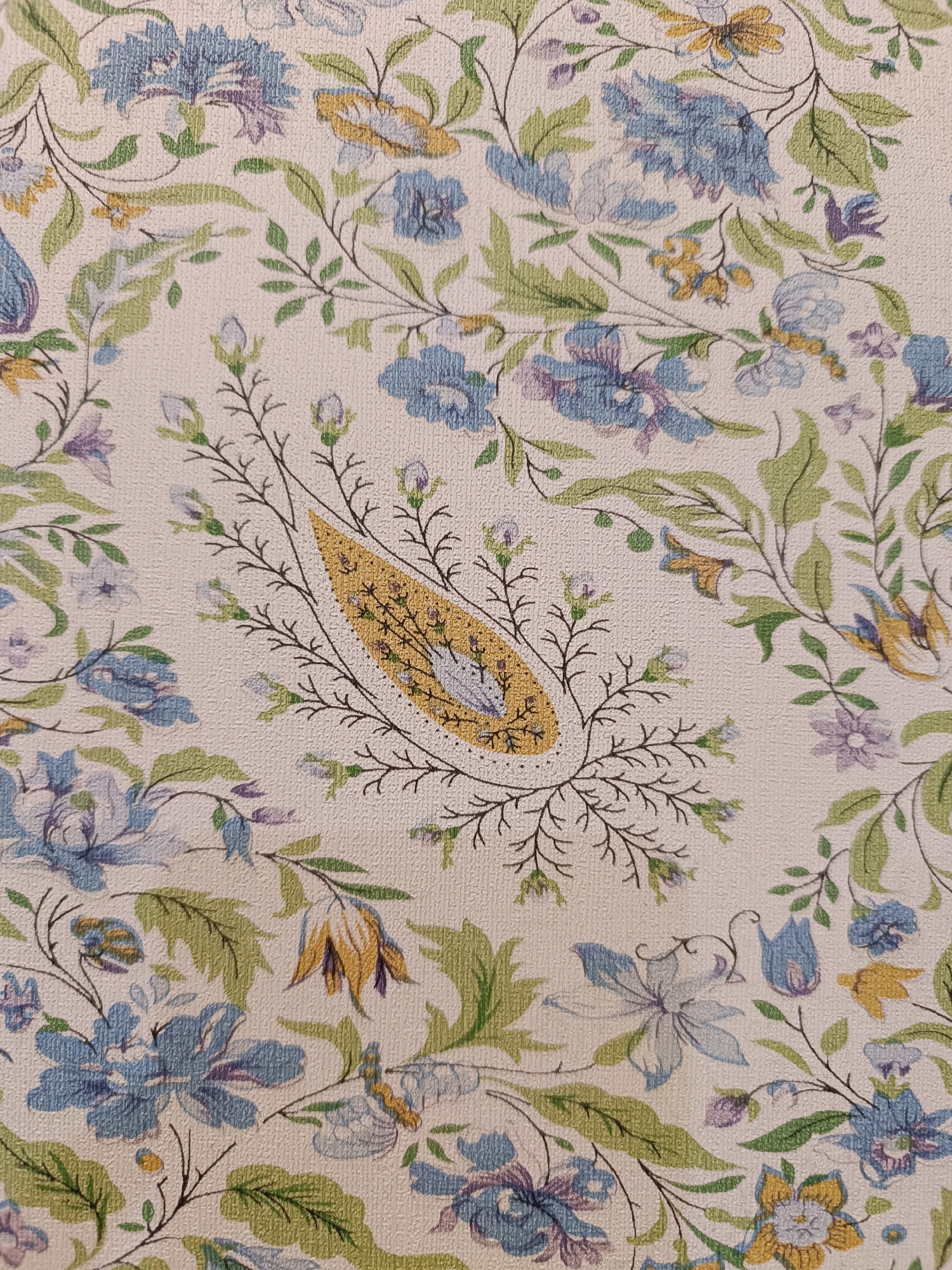 The image is a close-up, high-angle shot of a textured fabric with a floral pattern. The pattern features a profusion of flowers and leaves in shades of blue, purple, green, and yellow, set against a light, off-white background. The flowers are rendered in a somewhat stylized, painterly manner, with visible brushstrokes and a slight impressionistic quality. Some of the flowers are fully bloomed, while others are depicted as buds. Delicate, thin brown branches and stems weave through the floral elements, creating a sense of organic growth and interconnectedness. In the center of the image, a prominent paisley-shaped motif in yellow is outlined in dark brown and filled with intricate, dark brown linework, possibly depicting a bird or another small creature within its design. The overall impression is one of a vintage or traditional textile design, possibly wallpaper or upholstery fabric.
The prompt requests information about the location in the city of Srinagar, India. However, the image itself does not contain any visual cues or context that would allow for the identification of a specific location in Srinagar, India. The image solely depicts a fabric pattern.