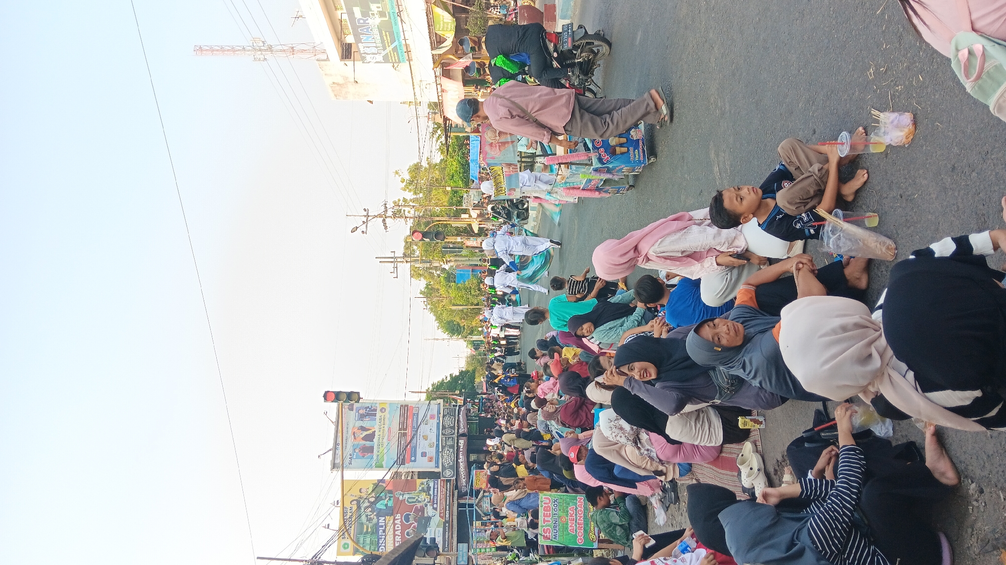 This is a vertical, eye-level, medium shot of a street scene in Tanjung Anom, Indonesia, during what appears to be a parade or procession. A large crowd of people, many wearing headscarves, are seated on the asphalt road on the left side of the image, facing right. They are observing a procession of individuals in white uniforms marching down the street, some carrying flags or banners. In the foreground, several children are also seated on the road. To the right of the street, there are buildings, traffic lights, and utility poles. Two ice cream carts are visible, one of which is being pushed by a person. A motorcycle is parked on the right side of the road. The overall atmosphere appears to be festive and communal.