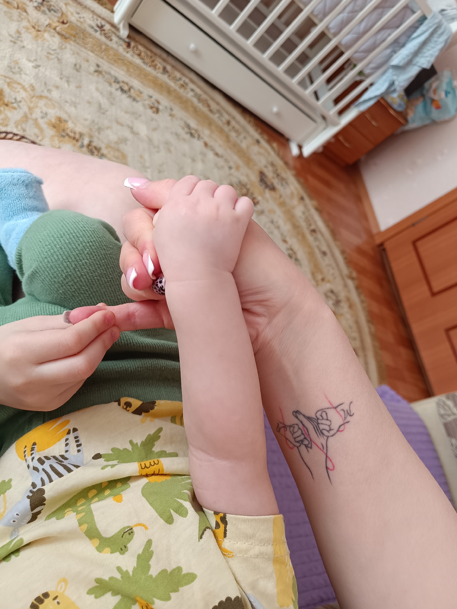 A close-up, high-angle photograph shows a fair-skinned adult holding the hand of a young child. The adult's left forearm is visible, bearing a black line tattoo with red accents, depicting hands intertwined with a red string. The adult has manicured nails with a French tip and a decorative nail on the thumb. The child's arm, wearing a green long-sleeved shirt with a yellow patterned bottom depicting animals and trees, holds the adult's index finger. The child also wears a blue sock.
A section of beige carpet with a pattern is in the background, along with a white crib and wooden furniture. The scene appears to be indoors. There is no information indicating the image's location as Tobolsk, Russia.