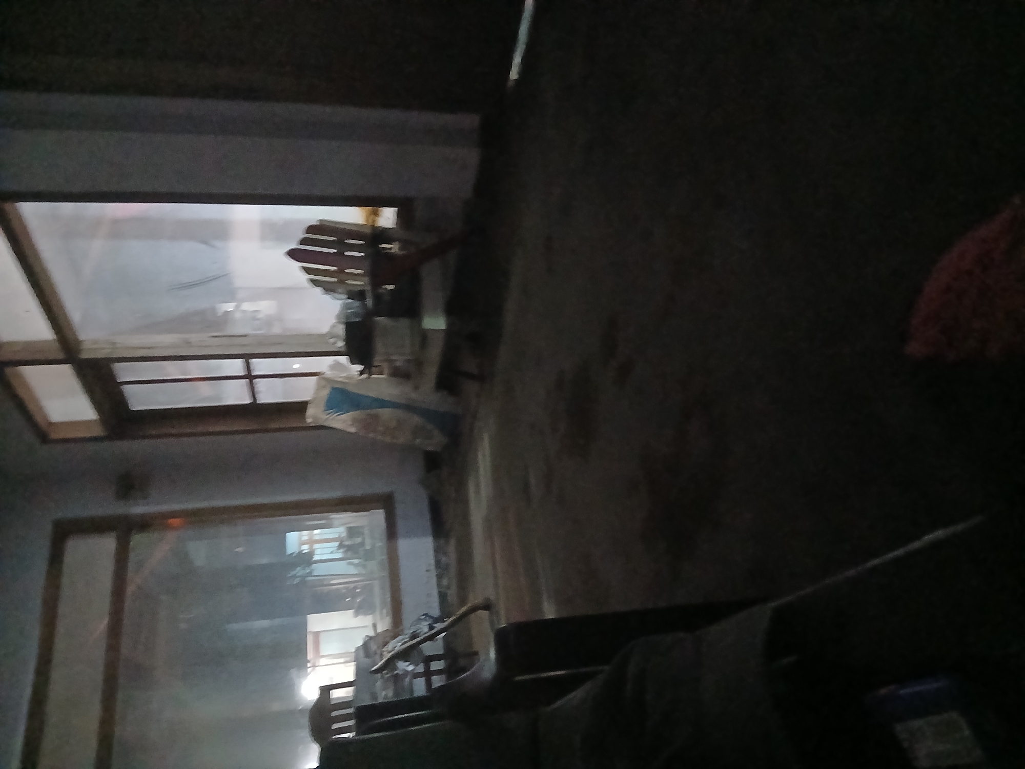 The image shows an indoor scene with large windows. The room appears to be sparsely furnished, with a wooden chair and a table visible. Outside the windows, there is a blurry view of what might be buildings or other structures. The lighting in the room is dim, suggesting it might be evening or nighttime. The floor appears to be covered in a rough material, possibly dirt or concrete. The overall impression is of an unfinished or disused space.