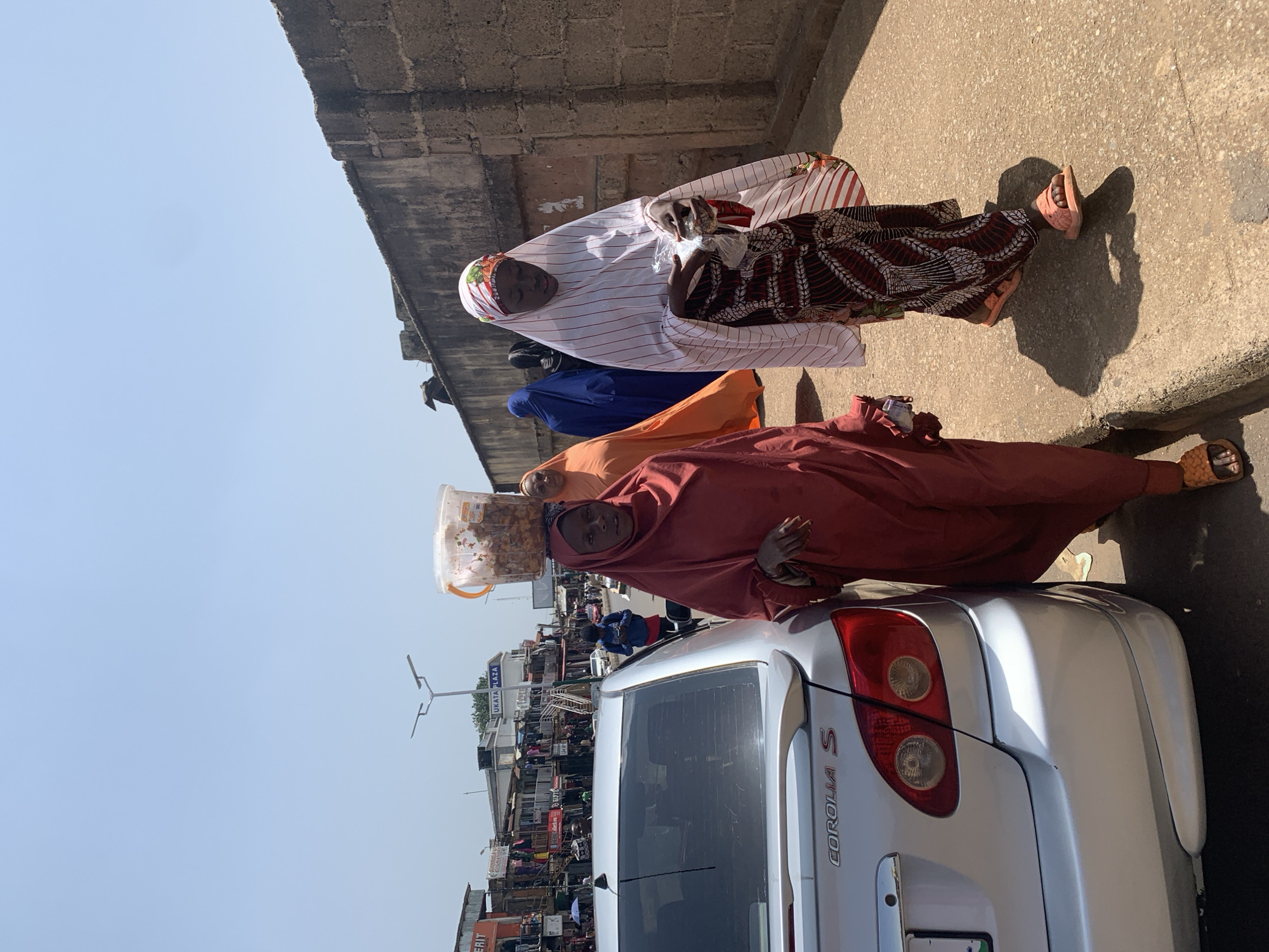 In Jos, Nigeria, on a clear, sunny day, a silver Toyota Corolla S sedan is parked on the right side of a paved road. A woman wearing a red hijab and a matching long dress walks beside it, carrying a clear plastic bucket filled with food items on her head. To her right, another woman in a white and red striped hijab and a patterned skirt walks towards the viewer, holding a clear plastic bag. Behind them, several other women are visible, some in colorful hijabs, and further in the background, a bustling marketplace with shops and people can be seen. The scene takes place along a textured concrete wall.