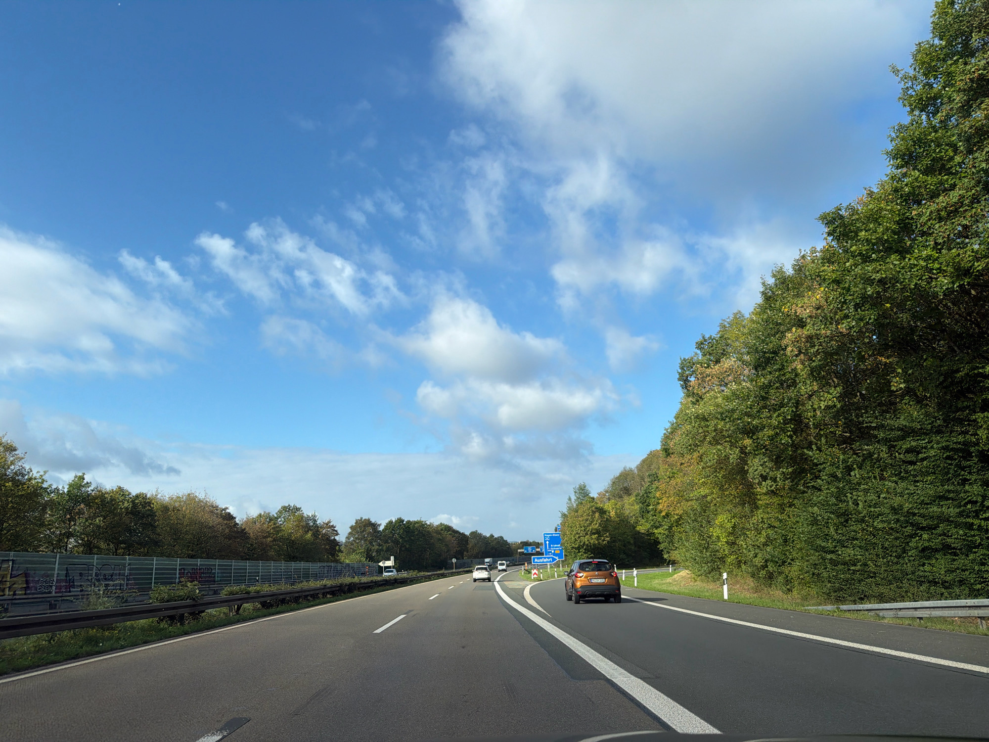 The image shows a highway in Germany with several cars driving on it. On the left side of the highway, there is a sound barrier with graffiti on it, followed by trees. On the right side of the highway, there are trees and a sign indicating an exit. The sky is blue with some white clouds. The image was taken from a moving vehicle. Based on the exit sign indicating "Hagen-Nord / Iserlohn-Letmathe", this scene is likely on the A45 autobahn near the border of Hagen and Iserlohn, Germany.