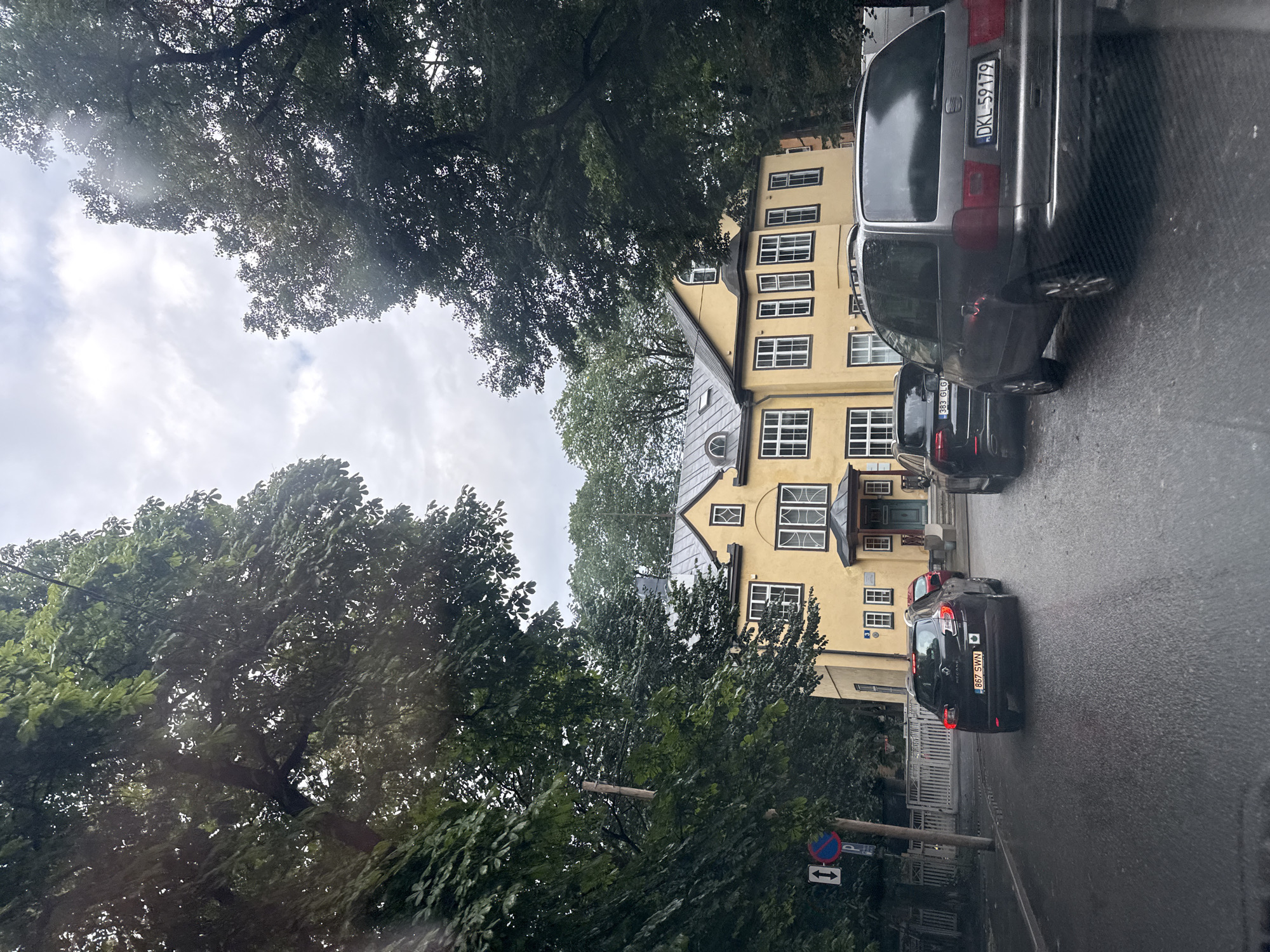 The image shows a street scene in Tallinn, Estonia. A yellow building with a gray roof is visible in the background, flanked by lush green trees. Several cars are parked and driving on the wet asphalt road. A black Renault hatchback with license plate "867 SWN" is in the foreground, while a gray minivan with license plate "DKL-59179" is to its right. Another black car with license plate "383 GLG" is parked behind the Renault. A traffic sign with an arrow and a red circle with a diagonal line is visible on the left side of the road. The sky is overcast.