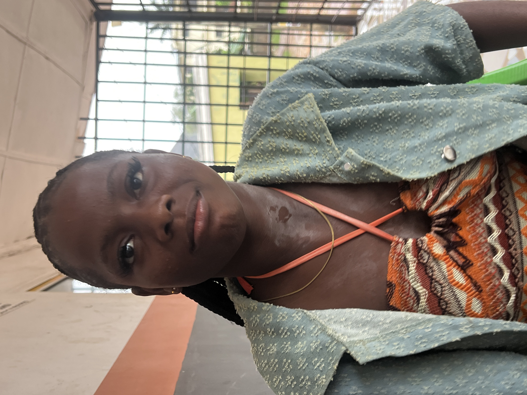 A medium shot captures a young Black woman with braided hair and dramatic false eyelashes looking directly at the camera. She is wearing a light blue textured button-up shirt over a patterned halter top in shades of orange, brown, and white. Two necklaces are visible: a thin gold chain and a thicker orange cord. Her skin appears slightly sweaty, particularly on her neck and chest. The background consists of a textured wall on the left, a metal grillwork fence, and a blurred yellow building with windows. The scene suggests an outdoor or semi-outdoor setting, possibly a veranda or sheltered walkway, consistent with a location in Nkpor, Nigeria. The lighting appears natural, indicating daylight.