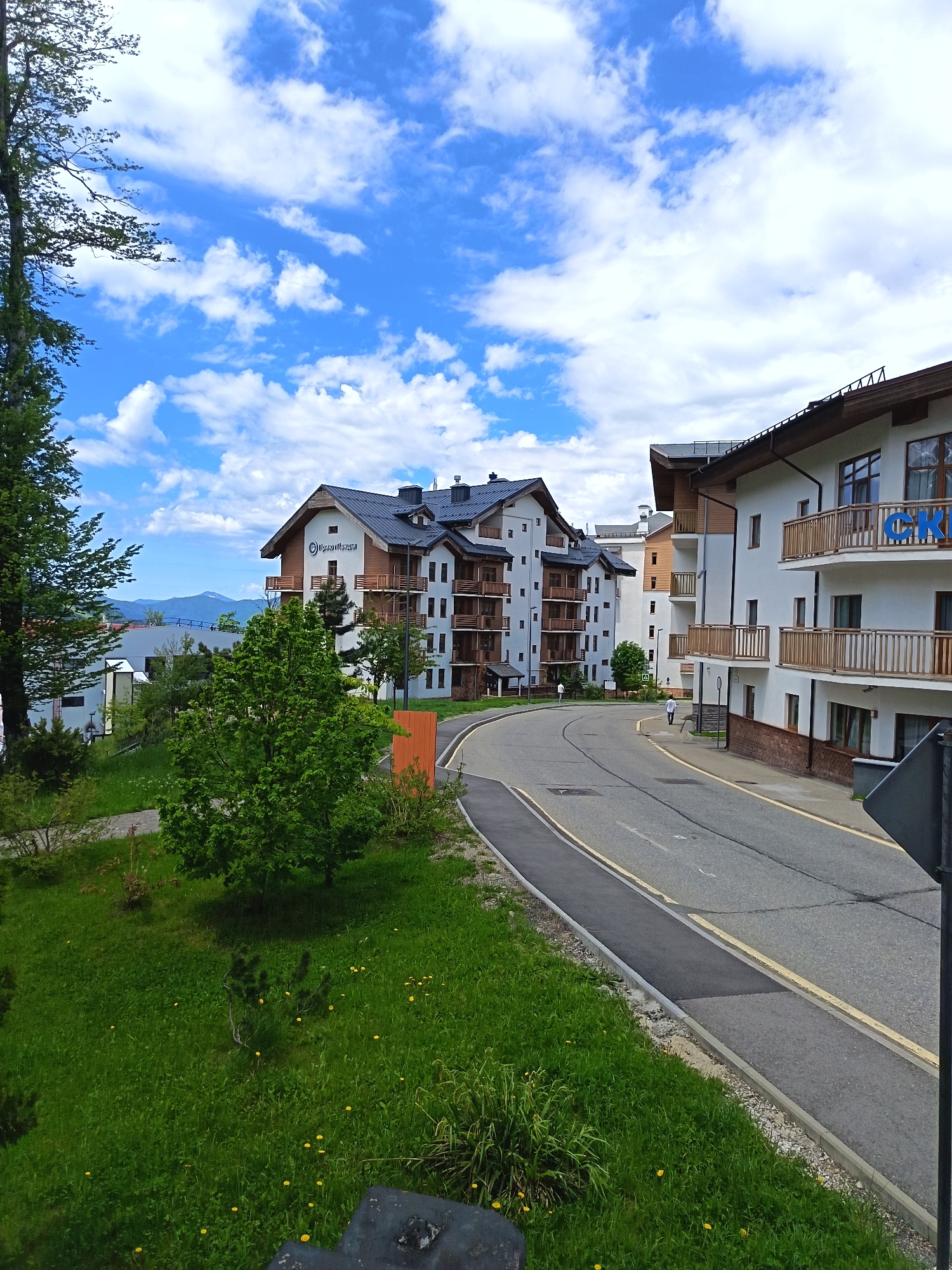 This image depicts a sunny daytime scene in Estosadok, Russia, showcasing a modern mountain resort or residential complex. A paved asphalt road, marked with double yellow center lines and single yellow lines delineating a pathway, curves gently through the frame.
On the left side of the road, a lush green grassy slope is visible, dotted with numerous small yellow dandelions. Several trees of varying sizes are interspersed within the greenery, including a tall, leafy tree on the far left and smaller, bushy trees closer to the road. In the background, beyond the buildings and trees on the left, distant mountains rise under a bright blue sky filled with scattered white, cumulus clouds.
Multi-story buildings with white facades, dark gabled roofs, and wooden balconies line both sides of the road. The architecture appears contemporary with traditional elements. On the prominent building to the left, the Cyrillic text "РозаПлаза" (Roza Plaza) is clearly visible on its upper facade. On a building on the right side of the road, the blue letters "CKI" are visible, likely part of the word "SKI." Two individuals can be seen walking in the distance on the road or sidewalk. The scene conveys a peaceful and well-maintained environment under pleasant weather conditions, likely late morning or early afternoon.
