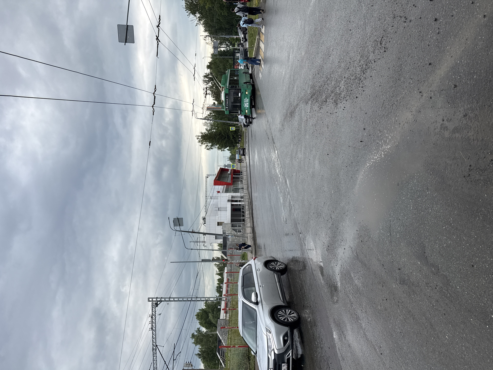 The image depicts a street scene in Kazan, Russia, likely during an overcast day due to the cloudy sky and wet asphalt. In the foreground, a silver SUV is parked on the right side of the road, its surface reflecting the diffused light. The road appears wet, with puddles visible, suggesting recent rain. Overhead, trolleybus wires crisscross the sky, indicating a public transportation route.
Further down the street, a green trolleybus is stopped, with several people crossing the road at a designated crosswalk. Some pedestrians are walking away from the trolleybus, while others are approaching it. A red building, possibly a structure under construction or a modern architectural piece, is visible in the background to the left. Further in the distance, trees line the street, and more buildings can be seen. The overall atmosphere suggests a typical urban environment, possibly a transit hub or a busy street.
The trolleybus displays Cyrillic text, and the license plate of the SUV also shows Cyrillic characters, consistent with a Russian location. The weather appears to be cool and damp. The scene is captured from a low angle, looking upwards towards the sky and across the street.