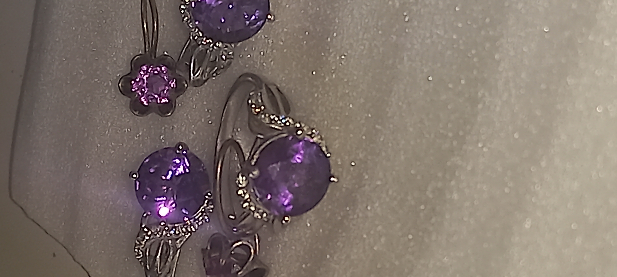 The image shows a close-up of amethyst jewelry, including rings and earrings, set against a light-colored, possibly textured surface. No people, actions, or location details are discernible.