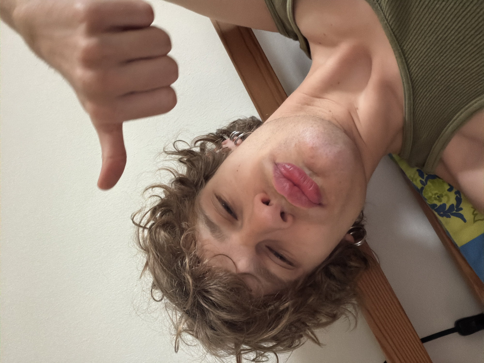The image is a selfie taken from a low angle, showing a person with light brown curly hair, fair skin, and wearing an olive green tank top. The person is giving a thumbs-down gesture with their left hand, which is positioned in the upper left corner of the frame. The person has multiple silver hoop earrings in their left ear. The background consists of a white wall on the left and a wooden frame with a patterned fabric on the right. The lighting appears to be natural. There is no information to indicate the city is Milan, Italy.