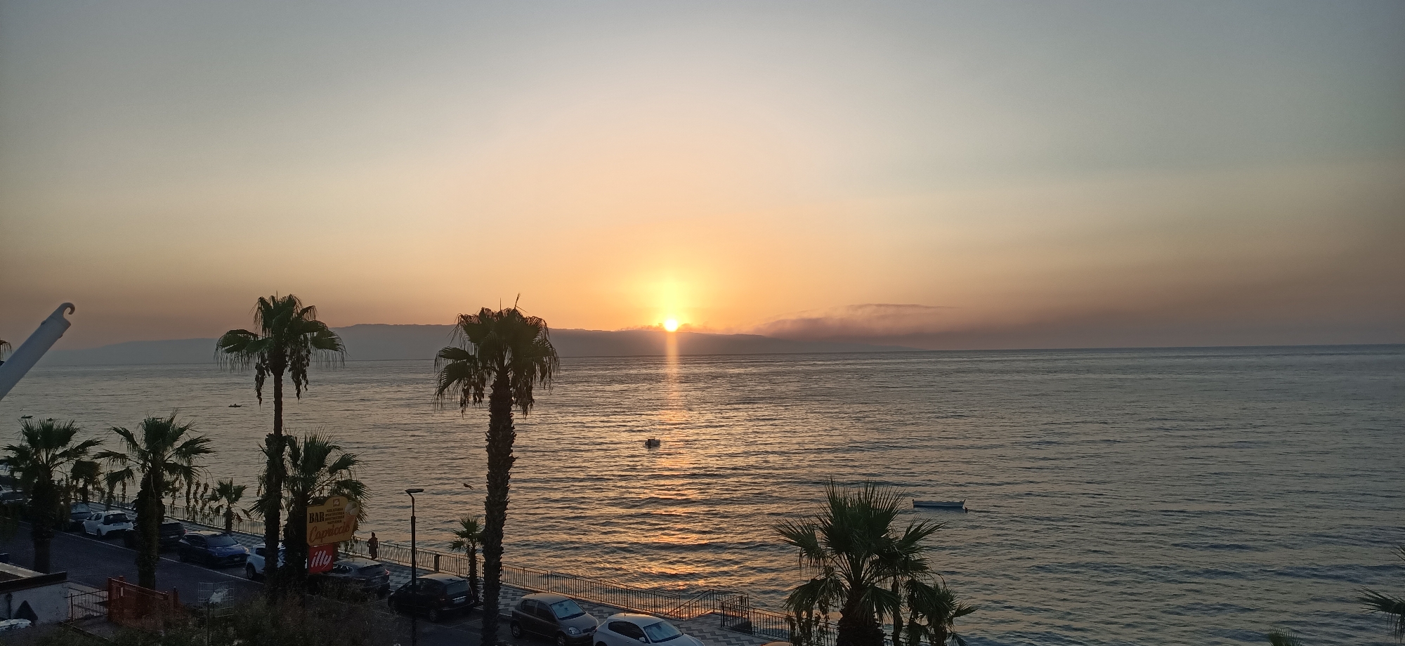 The image depicts a seaside scene at sunset in an unknown city. The sun, partially obscured by clouds, is setting over the ocean, casting a warm glow on the water and the sky. The sky transitions from a pale yellow near the horizon to a soft blue at the top. In the foreground, several palm trees line a waterfront promenade. A road with parked cars runs alongside the promenade. The sea is calm with gentle ripples reflecting the sunset. A few small boats are visible on the water. The overall atmosphere is serene and peaceful.