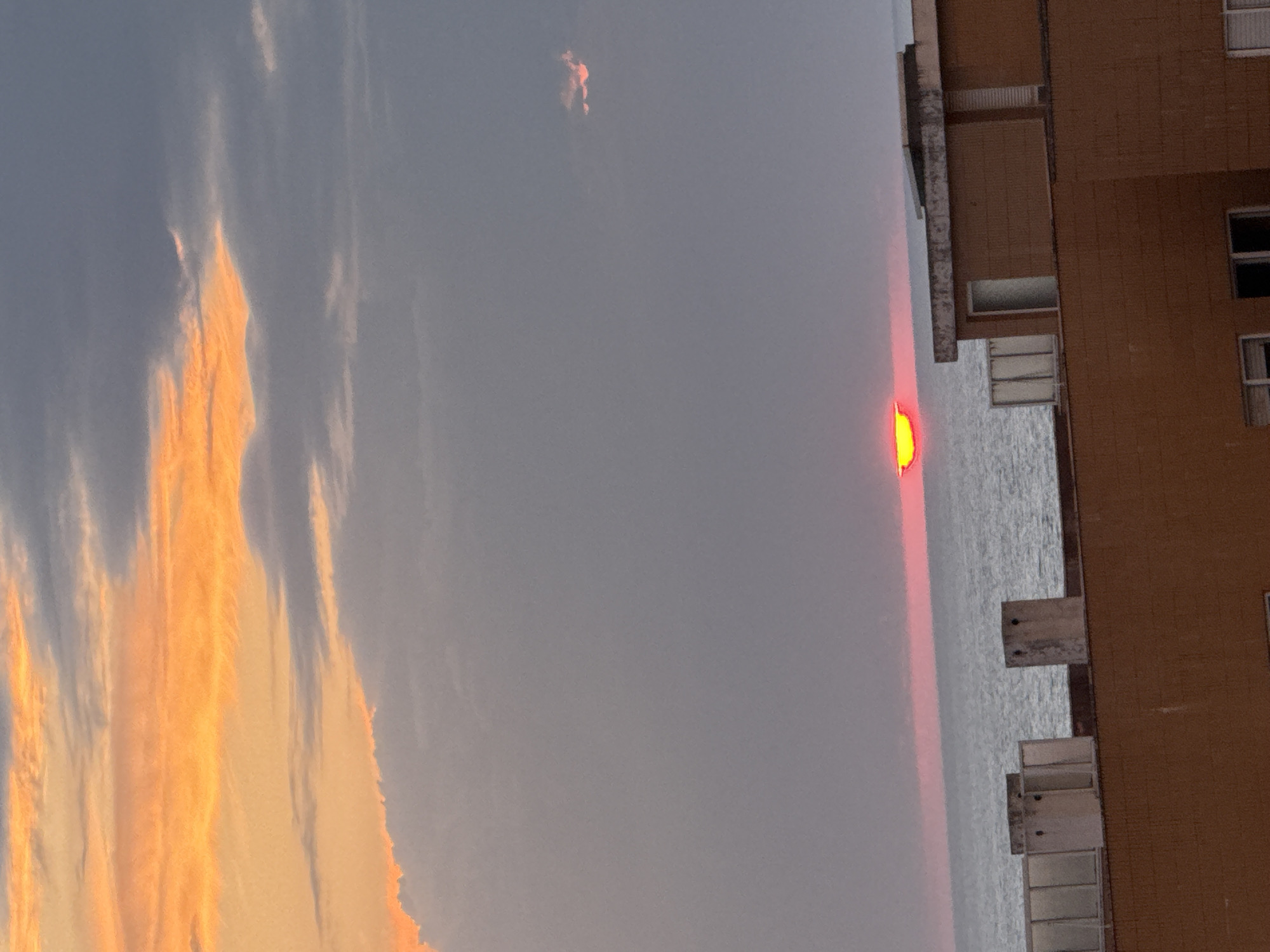 The image depicts a vibrant sunset over a body of water, likely the ocean or a large river, viewed from the perspective of an adjacent building. In the center, a bright, flattened, and intensely yellow-orange sun is visible just above the water's horizon, casting a distinct band of deep red light around it. The sky above transitions from a muted grey-blue to a brilliant display of orange and yellow, particularly on the left side, where elongated, wispy clouds are dramatically illuminated by the setting sun. A smaller, isolated cloud with an orange glow is also visible higher in the sky.
To the right, a multi-story building with a brown, possibly tiled, facade stands prominently. It features numerous rectangular windows and concrete architectural elements, which appear to be balconies or ledges extending towards the water. The surface of the water below the sun is rippled and reflects a shimmering grey light. The scene captures the golden hour, indicating either sunrise or more likely sunset, with clear skies apart from the illuminated clouds. The location context suggests this serene view is from Vila Nova de Gaia, Portugal. There is no visible text in the image.