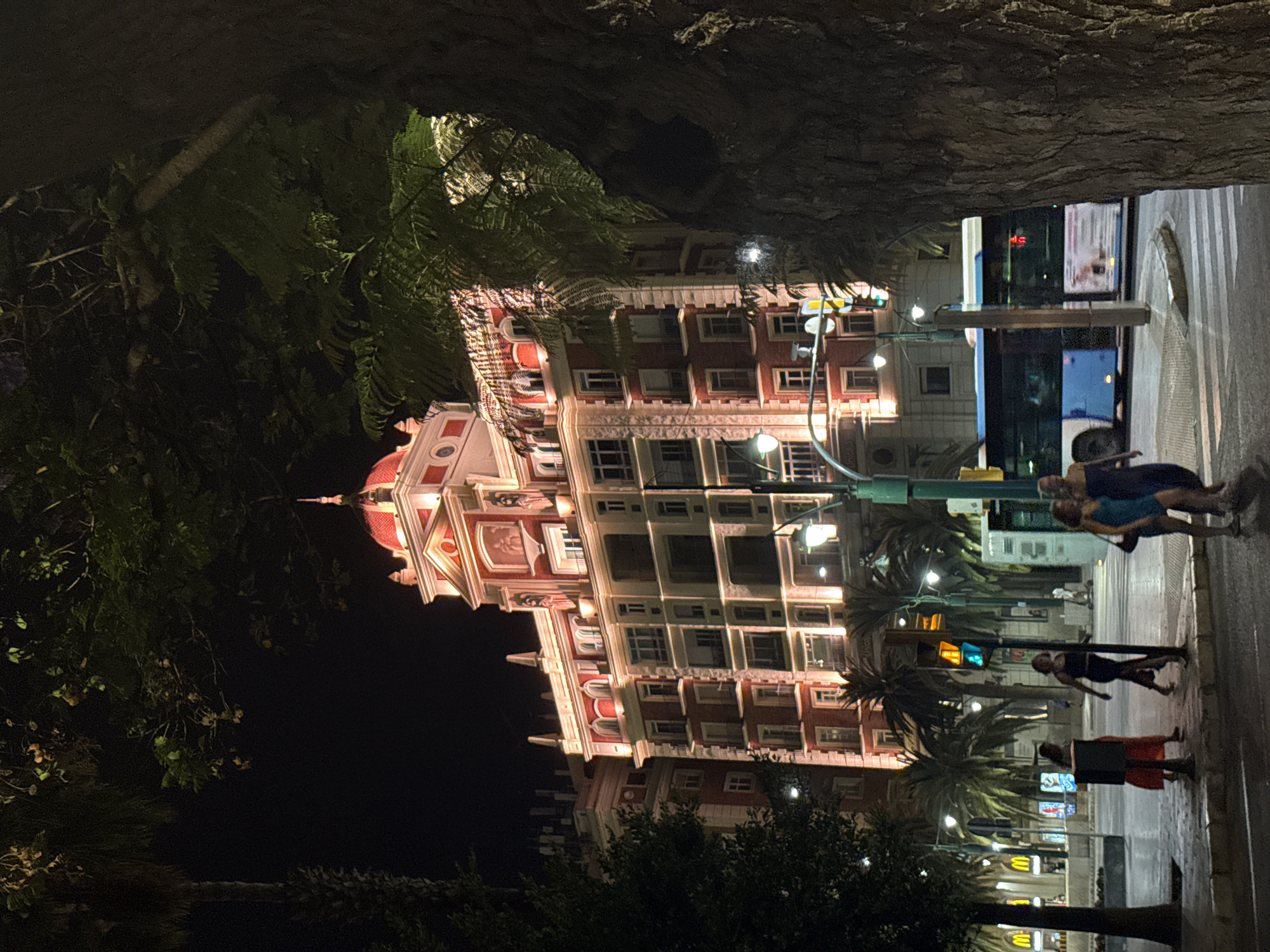 The image captures a nighttime scene on a street in Málaga, Spain, with a large, ornate building in the background. The building is red with white accents, featuring multiple floors with numerous windows and decorative elements, including arched windows and what appears to be a dome with a spire. The building is illuminated, casting a warm glow on its facade.
In the foreground, several people are visible walking on the sidewalk. Some are grouped together, while others are walking individually. The lighting suggests it is nighttime, and the street appears to be wet, possibly from recent rain, which is reflected in the light. Tropical palm trees are scattered throughout the scene, adding to the Mediterranean atmosphere. A bus is seen in the background, and traffic lights are also visible.
The overall mood of the image is lively, with the illuminated building and the presence of people suggesting a vibrant urban setting after dark. The composition, with the dark, textured foreground element framing the illuminated building and street, creates a sense of depth and draws the viewer's eye to the main subjects. There is no visible text in the image.
