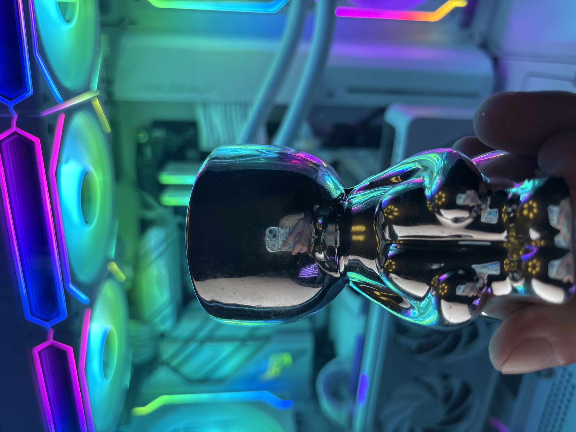 A person is holding a shiny, metallic figurine in front of a computer with RGB lighting. The figurine appears to be a stylized bear or character, with a reflective surface that mirrors the colorful lights of the computer components. The background is filled with the vibrant glow of RGB LED strips and fan lighting, predominantly in shades of blue, green, and purple. The computer case itself is partially visible, with white and gray elements. The lighting creates a futuristic and high-tech atmosphere. The focus is on the figurine, with the background slightly blurred.